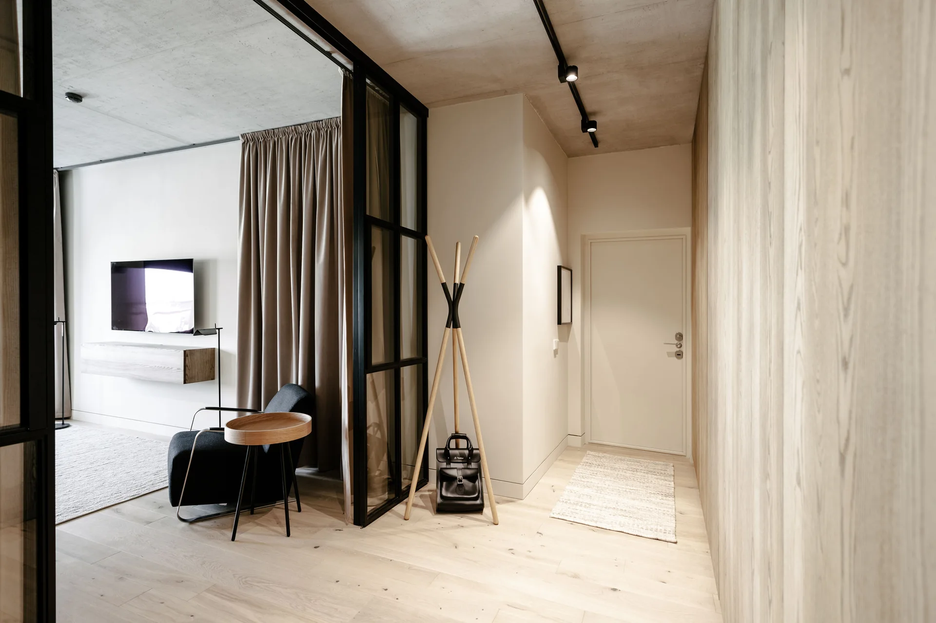 A minimalist entryway with a light wood floor and wall paneling, a wooden tripod coat rack, and a black-framed glass partition leading into a sitting area.