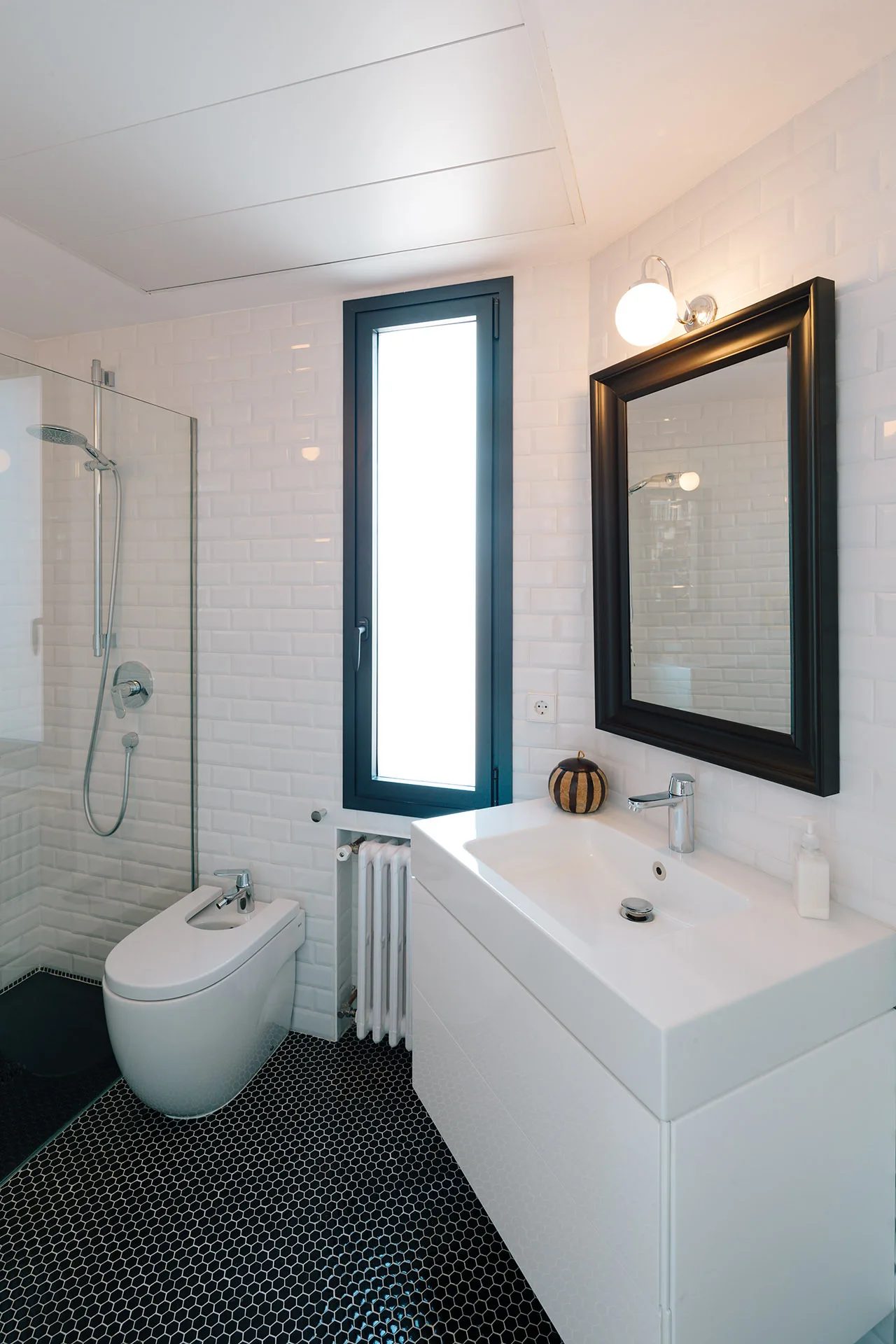 A modern bathroom features a white vanity, a bidet, and a glass shower next to white subway tiled walls and black hexagonal floor tiles.