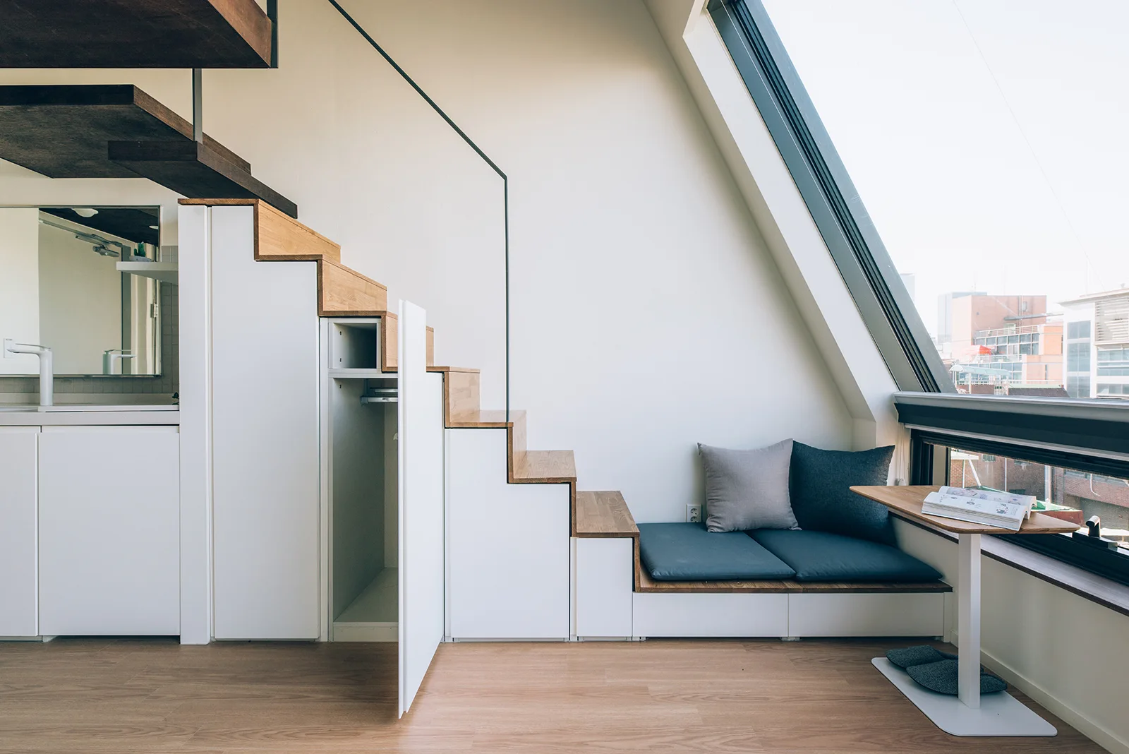 A modern wooden staircase features integrated white storage cabinets underneath and a blue cushioned bench next to a large sloped window.