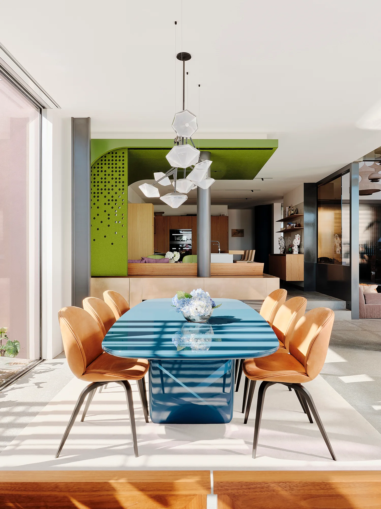 A glossy blue dining table is surrounded by tan leather chairs under a geometric glass pendant light in a modern open plan space.