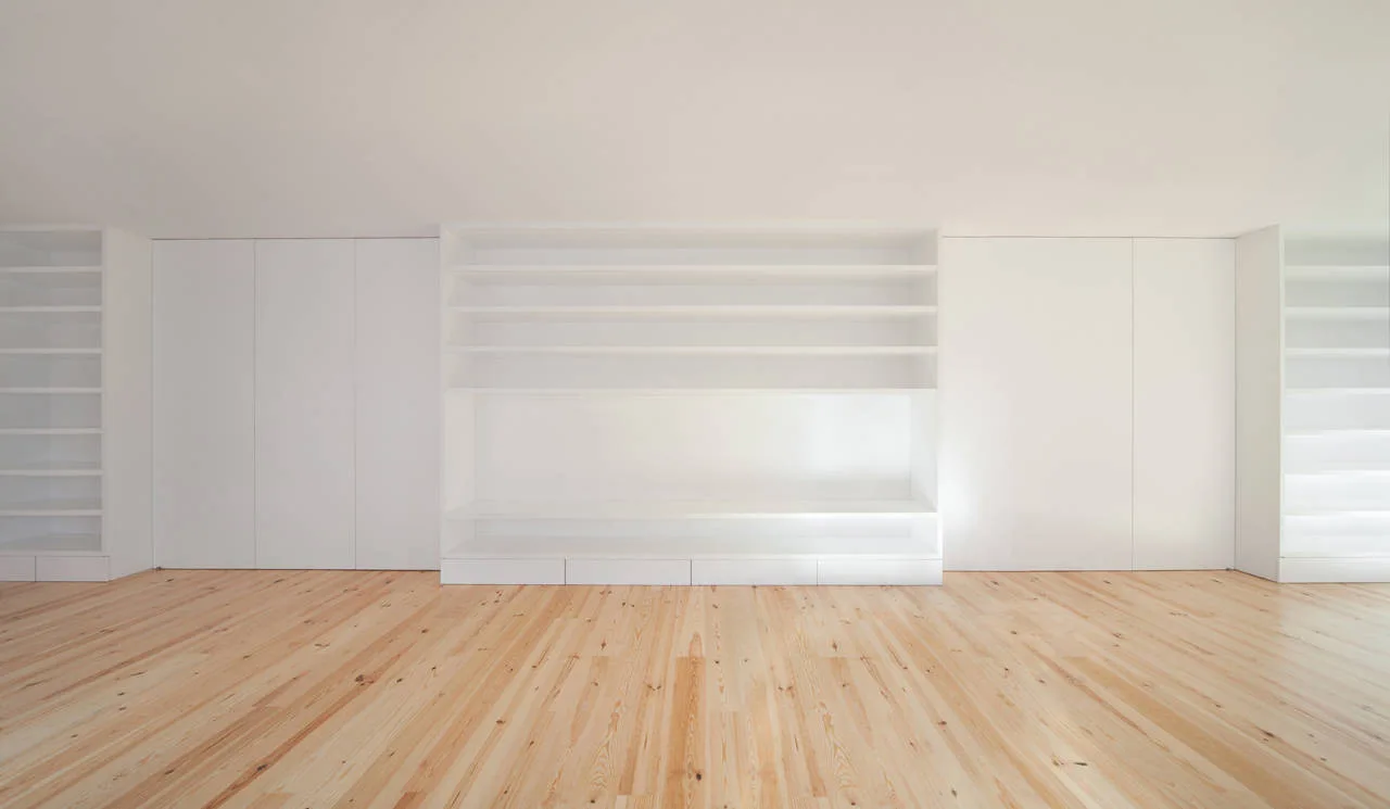 An empty minimalist room featuring a wall of custom white built-in storage cabinets, open shelves, and light natural pine wood plank flooring.