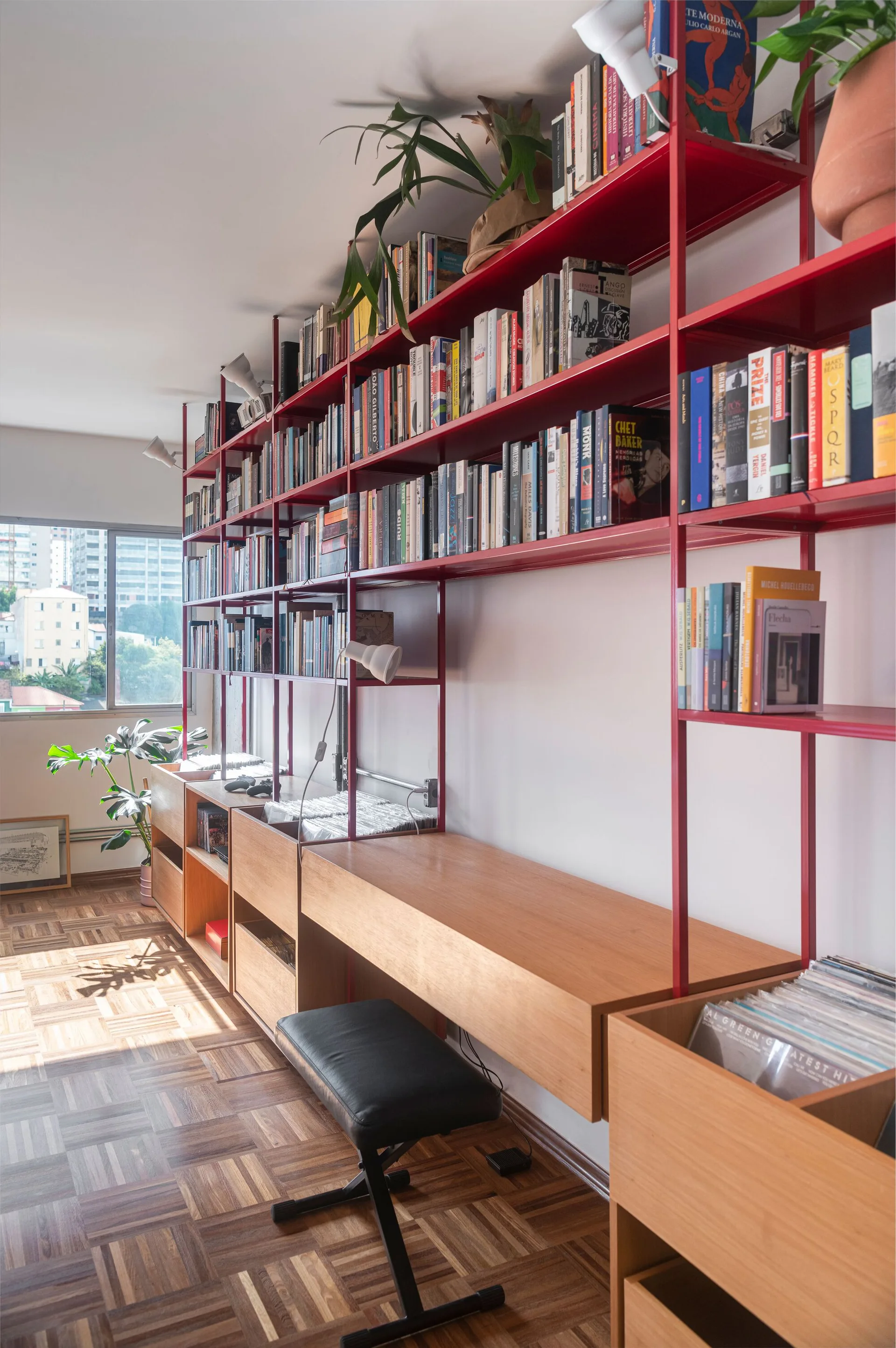 A home office features a red metal modular shelving unit with integrated wood desk surfaces and records next to a parquet wood floor.
