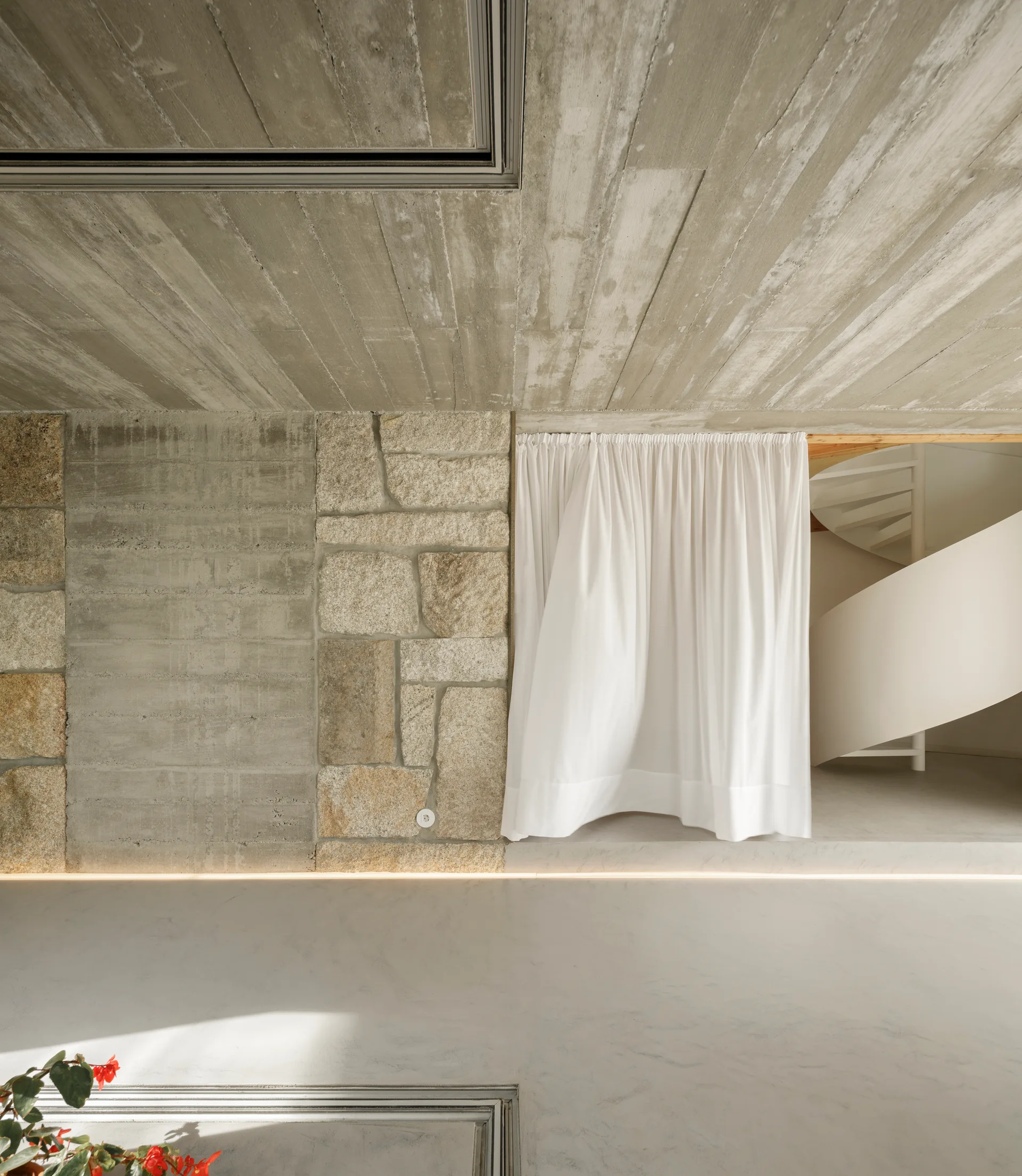 An interior space featuring a board-formed concrete ceiling, a mixed stone and concrete wall, and a white curtain revealing a spiral staircase.