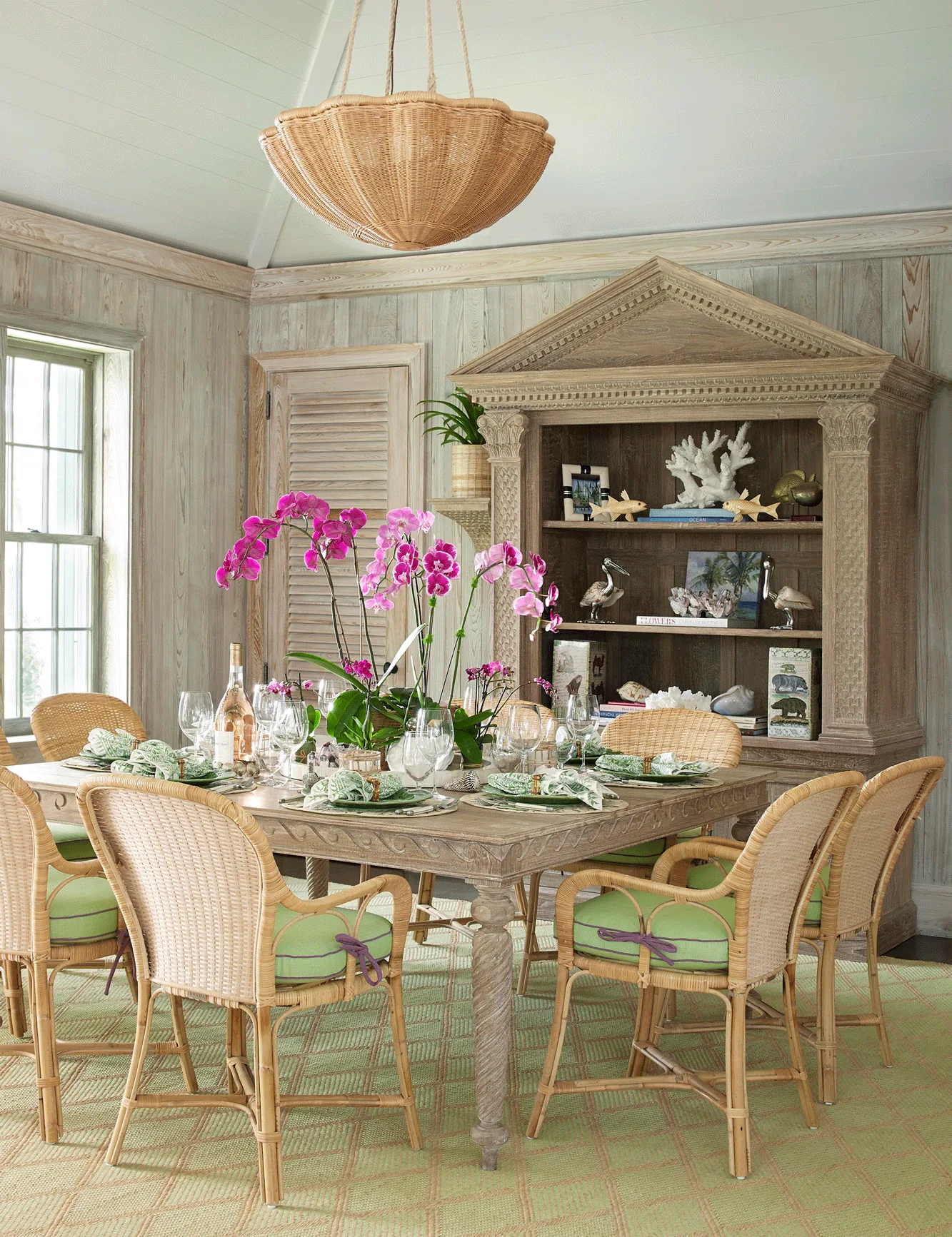 A coastal dining room featuring a weathered wood table, wicker chairs with green cushions, a large display hutch, and a woven pendant light.