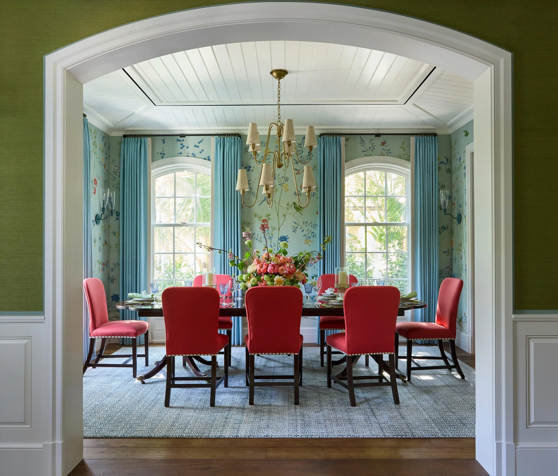A formal dining room framed by a white archway features bright red chairs around a dark wood table beneath blue botanical wallpaper.