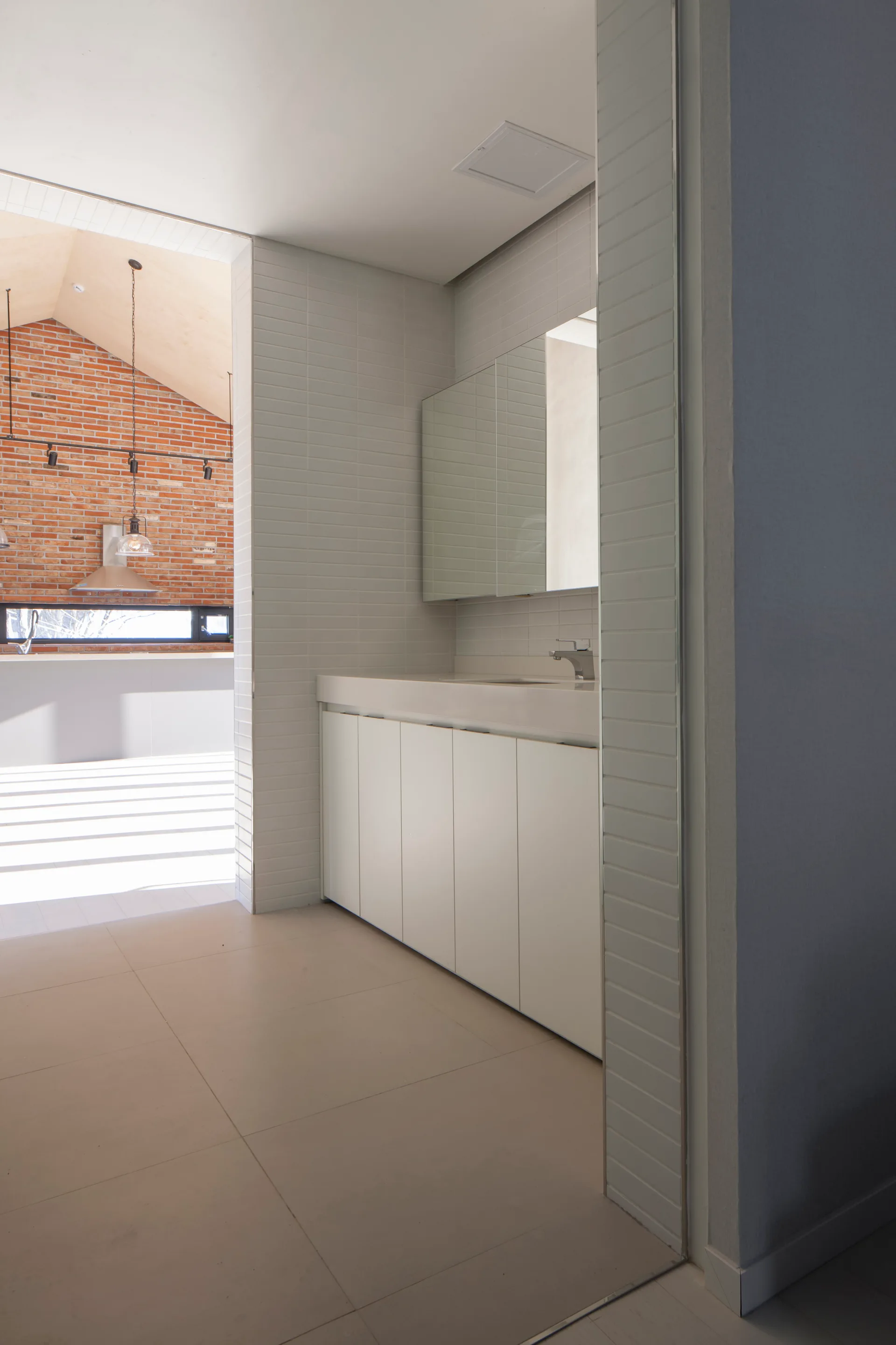 A minimalist white bathroom vanity with a large mirrored cabinet and white subway tile walls offers a view into an adjacent room with a red brick wall.