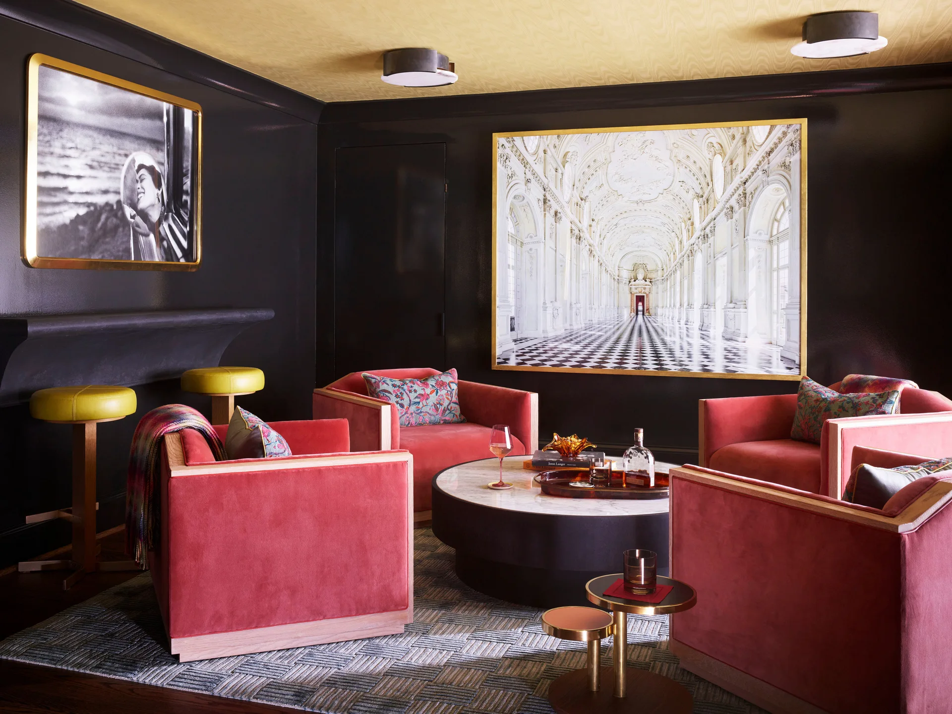 Four red velvet armchairs surround a round marble coffee table in a moody room with black walls, a gold ceiling, and framed art.