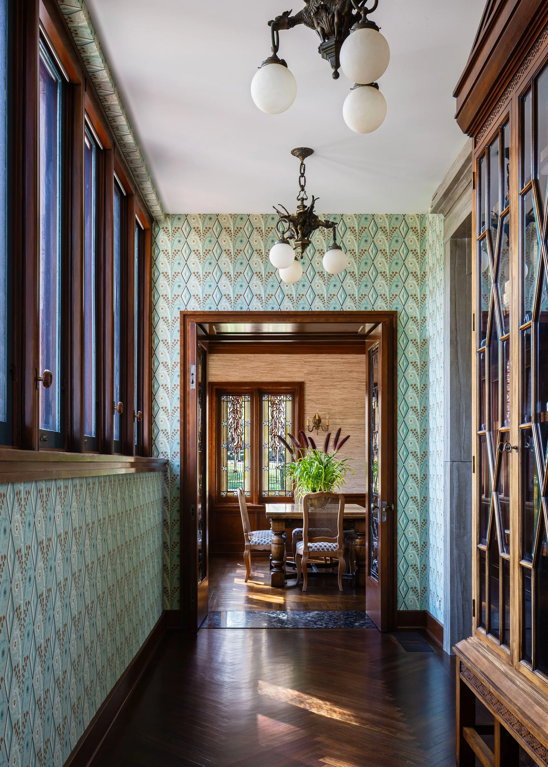 A hallway featuring blue patterned wallpaper, herringbone wood floors, and wood-framed windows leads to a dining room with a wooden table and stained glass.