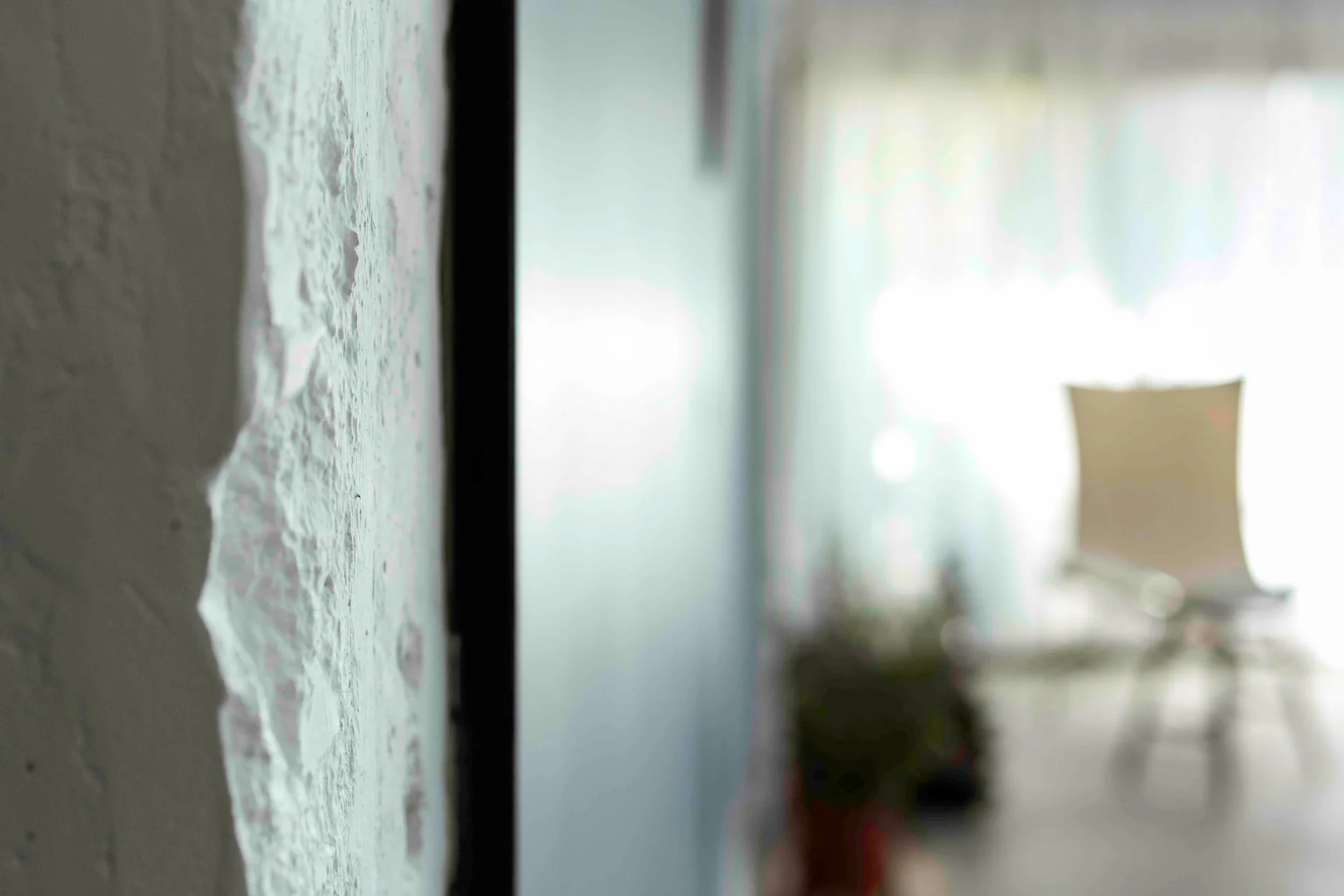 Close-up of a rough, textured white plaster wall in the foreground with a blurred silhouette of a chair and window in the background.