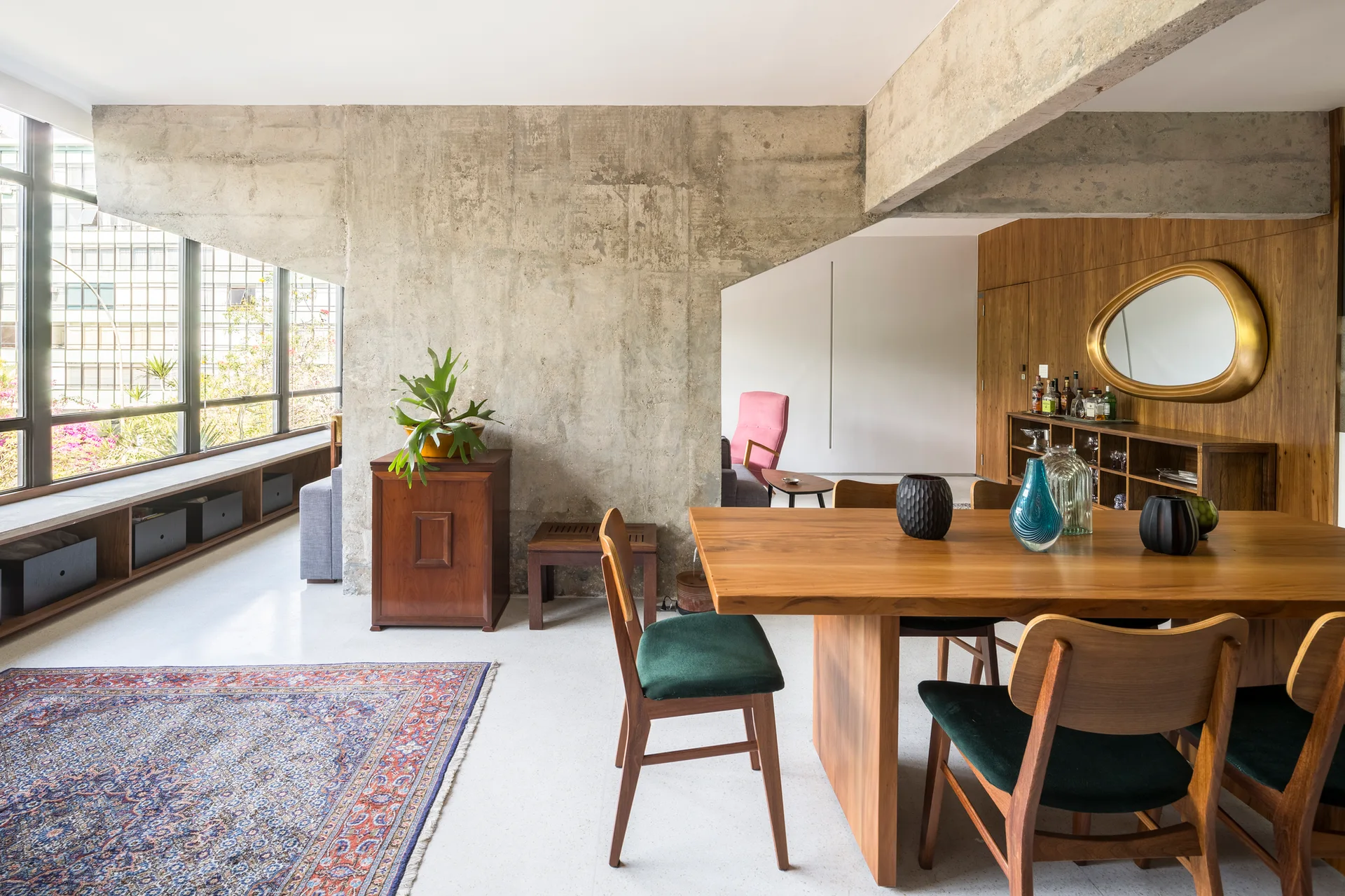 A wooden dining table with green velvet chairs sits near a massive exposed concrete pillar and a wood-paneled wall accented by an asymmetrical mirror.