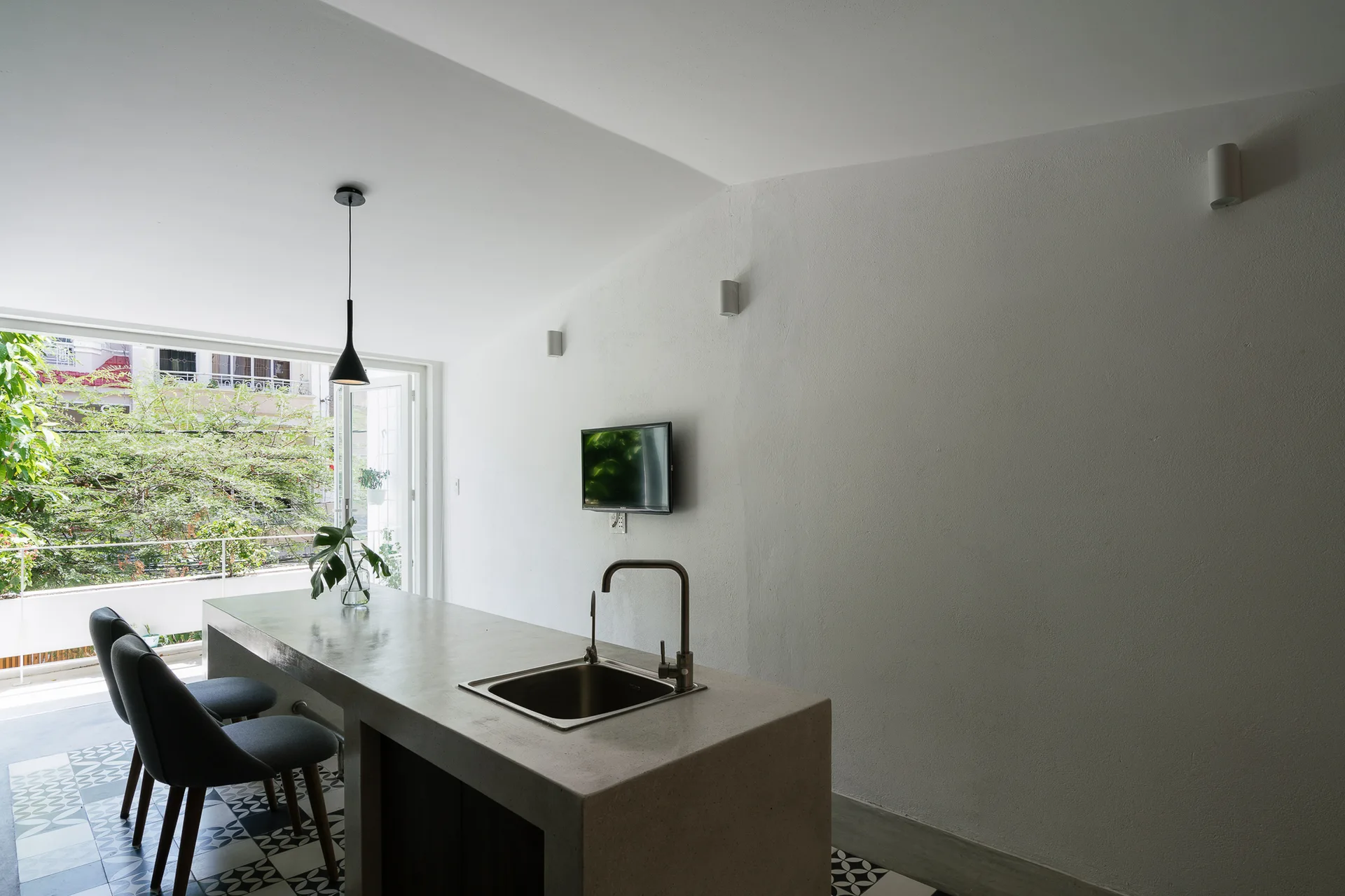 A minimalist kitchen features a solid gray concrete island with an integrated sink, dark bar stools, and bold black-and-white patterned floor tiles.
