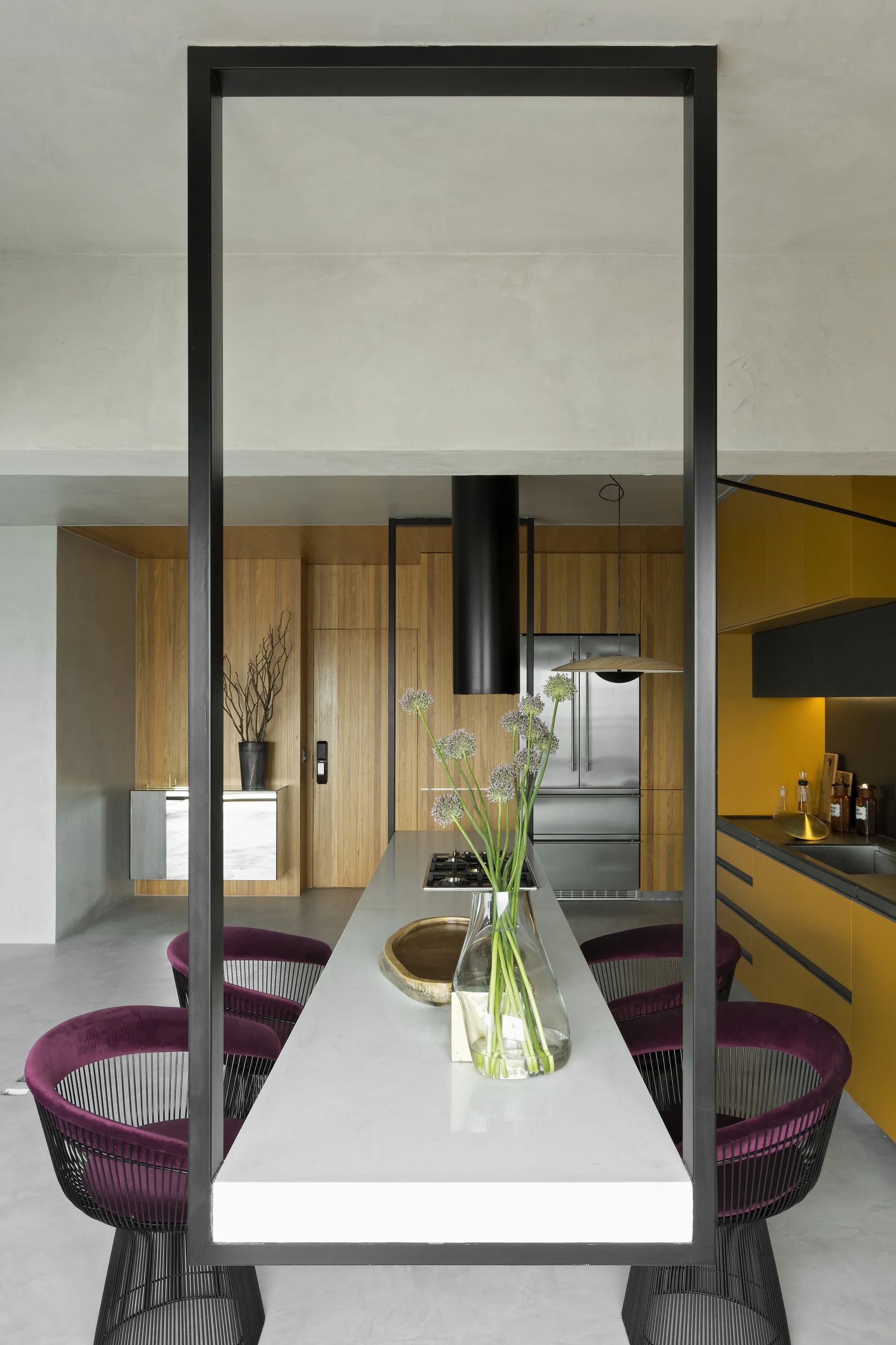 A modern kitchen featuring a long grey island with a black metal frame, purple velvet chairs, and mustard yellow cabinetry.