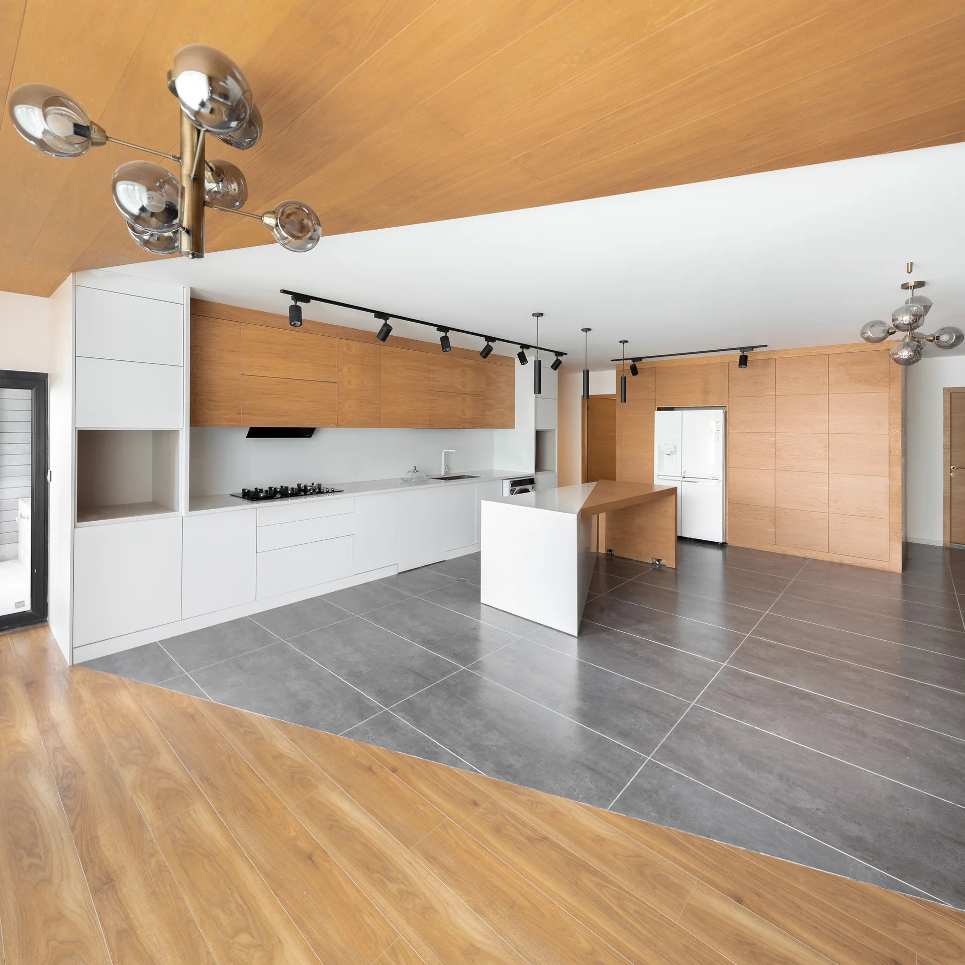 A modern kitchen with wood and white cabinetry, a white island with a wood dining extension, matte gray floor tiles, and an angled wood ceiling.