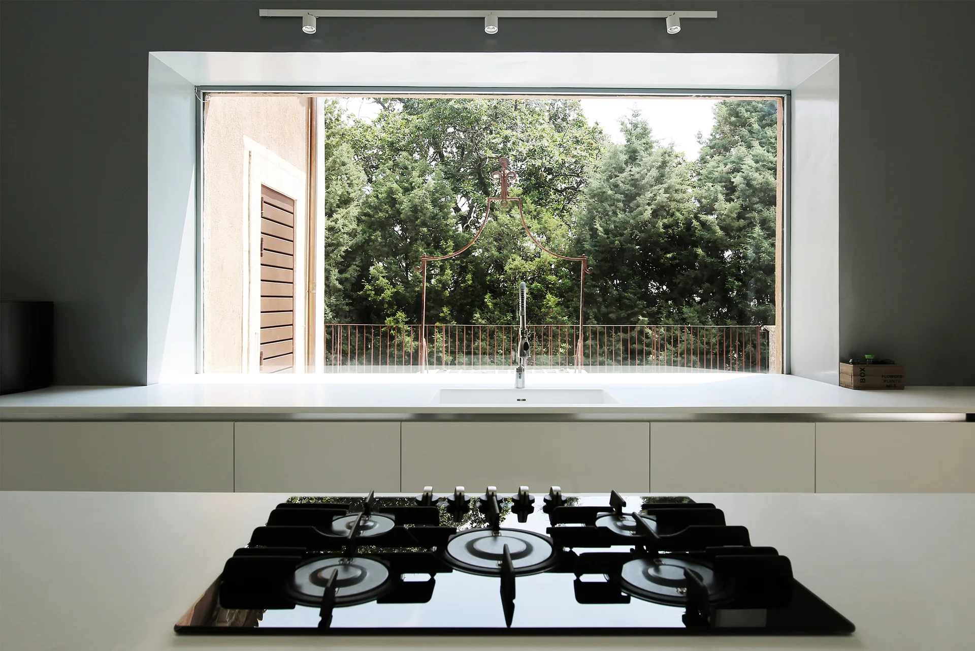 A modern minimalist kitchen features a white countertop with a gas cooktop in the foreground and a wide window framing lush green trees over the sink.