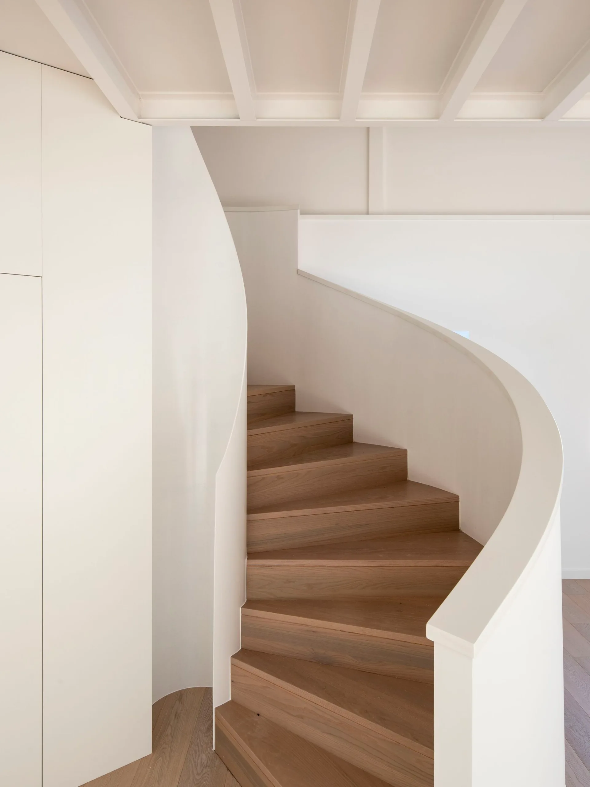 A curved staircase with warm wood treads is enclosed by smooth white plaster balustrades beneath a white-painted beamed ceiling.