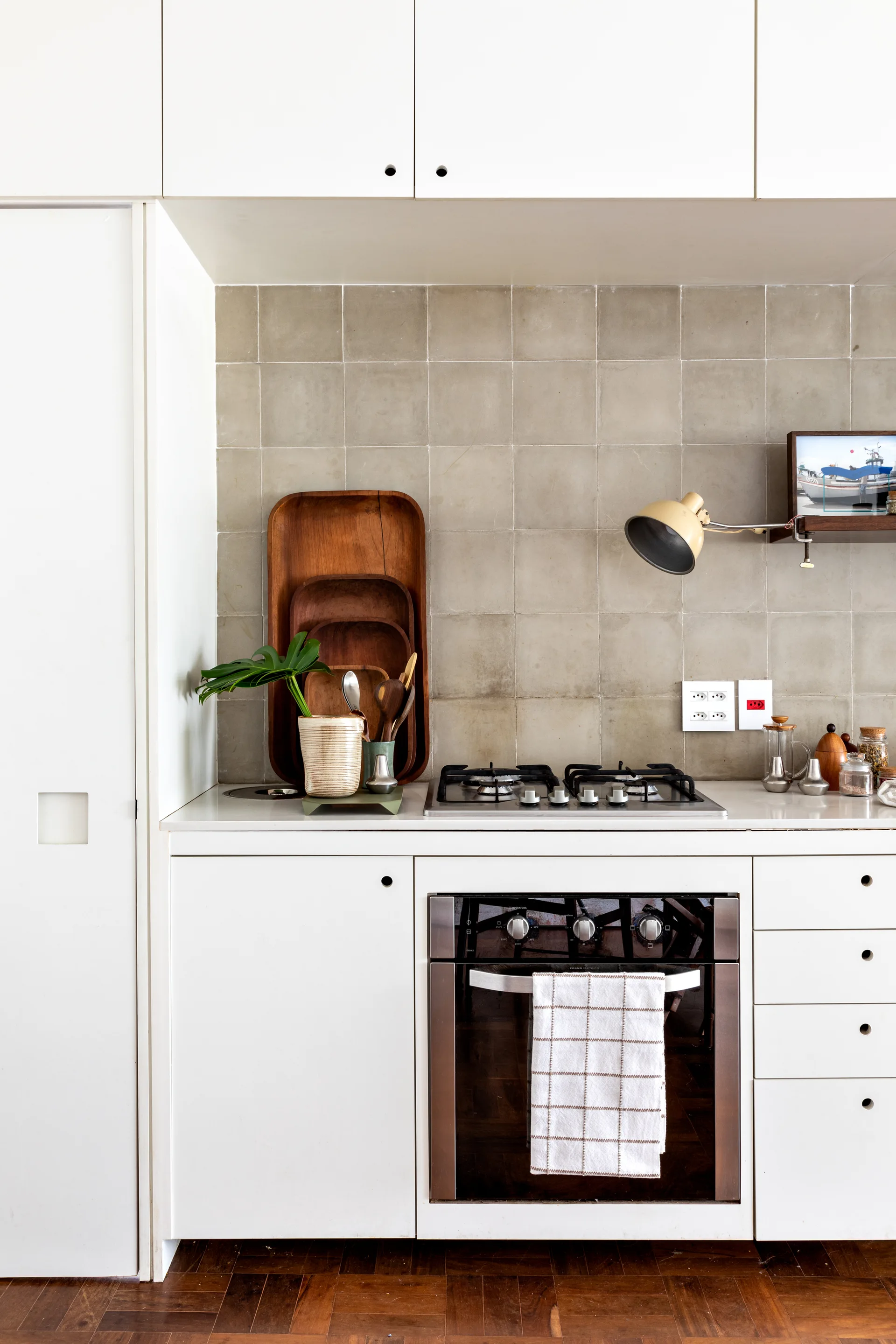 A modern kitchen featuring minimalist white cabinets, a grey square-tiled backsplash, a gas stovetop, and nested wooden trays on the countertop.