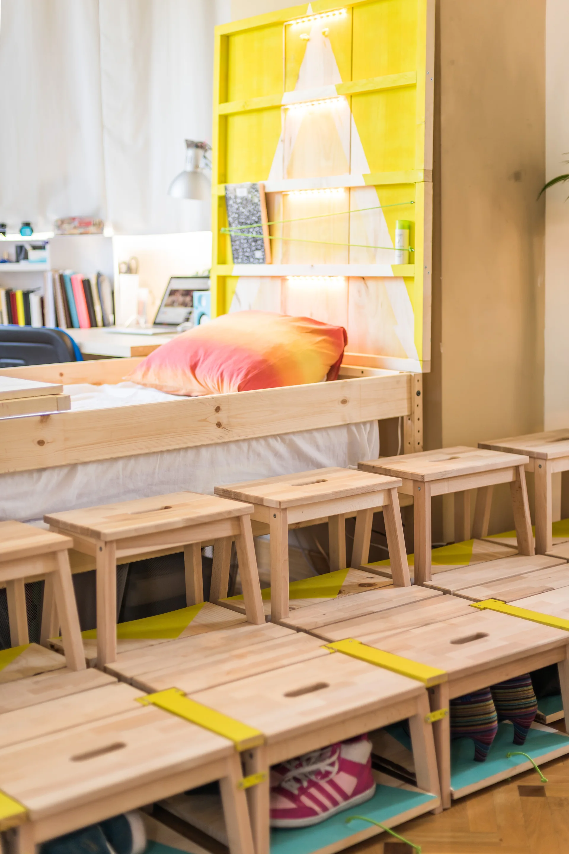 A compact bedroom features an elevated wooden bed frame, a bright yellow headboard with integrated lights, and a row of small wooden stools.