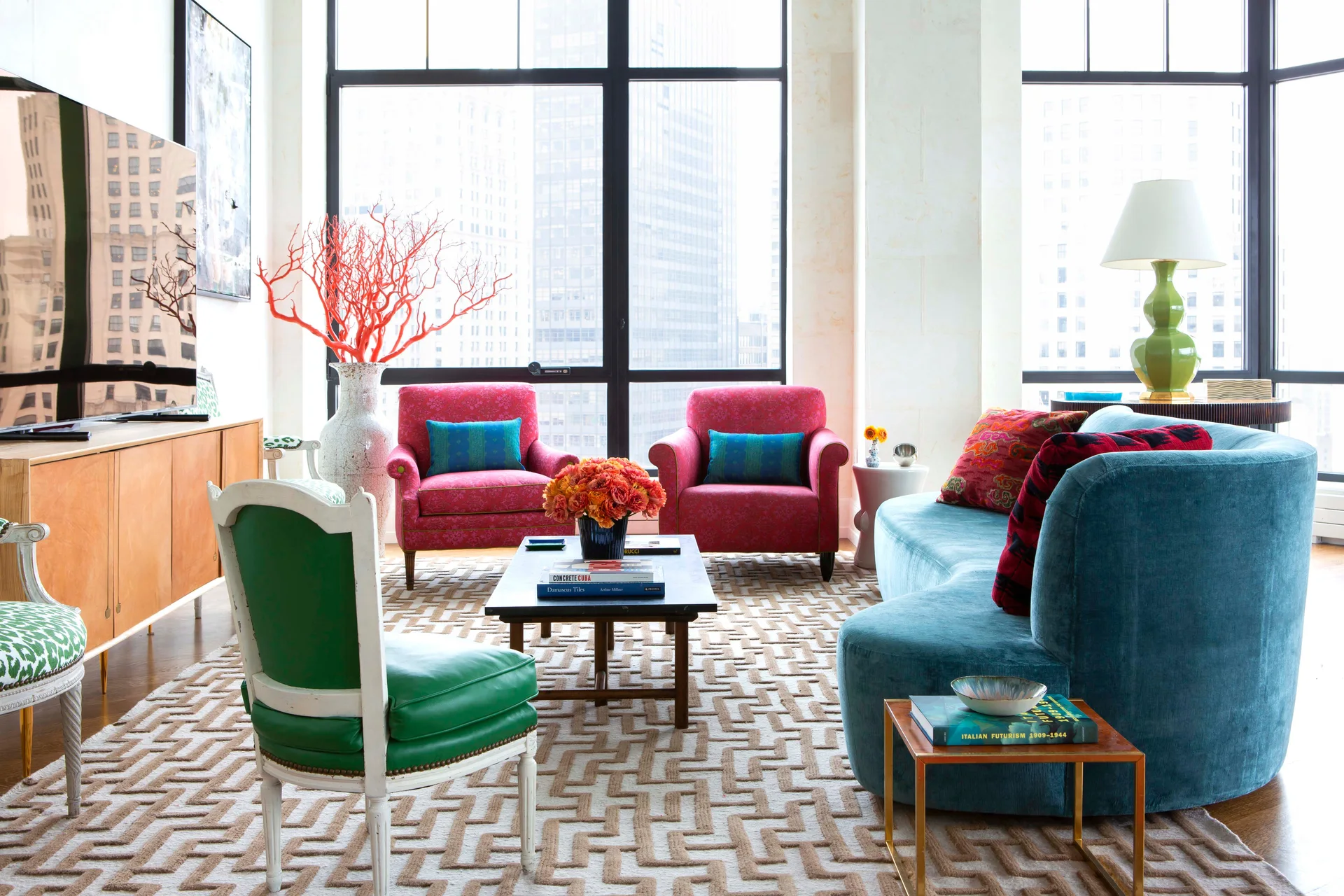 A bright high-rise living room features a curved teal sofa, two pink armchairs, a geometric rug, and large windows overlooking city buildings.