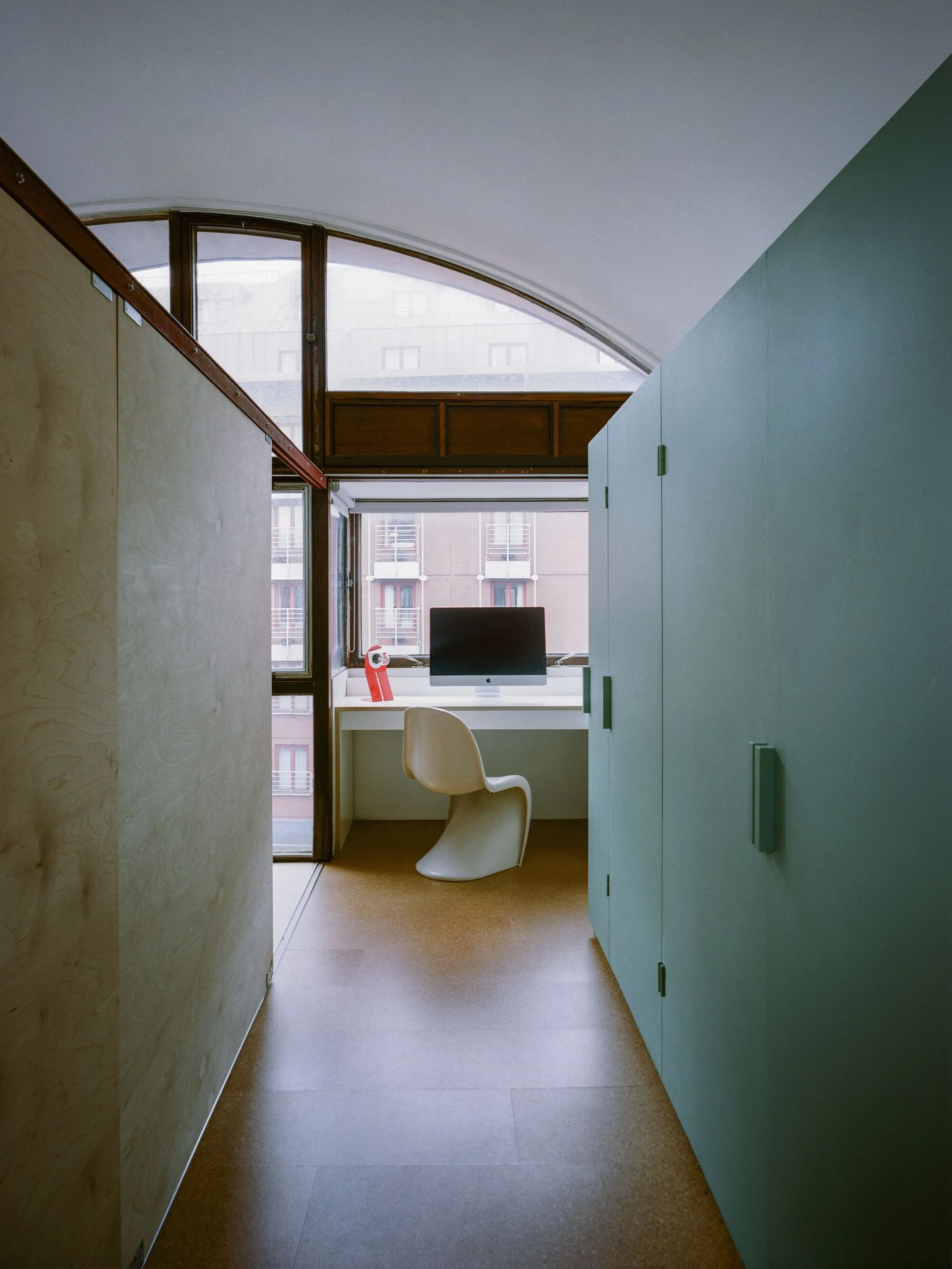 A narrow home office at the end of a corridor features a white desk, a Panton chair, and a sage green storage cabinet under an arched window.