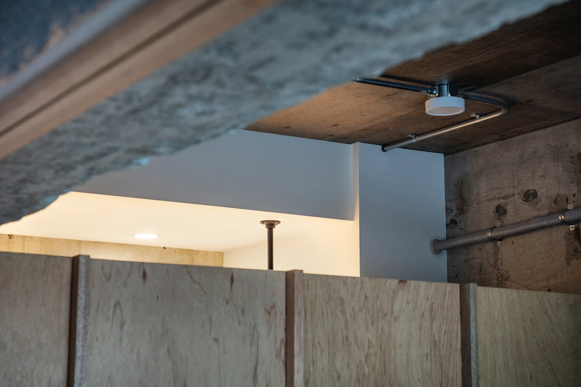 Raw concrete ceiling with exposed metal conduits and a surface-mounted light above warm plywood cabinetry panels.
