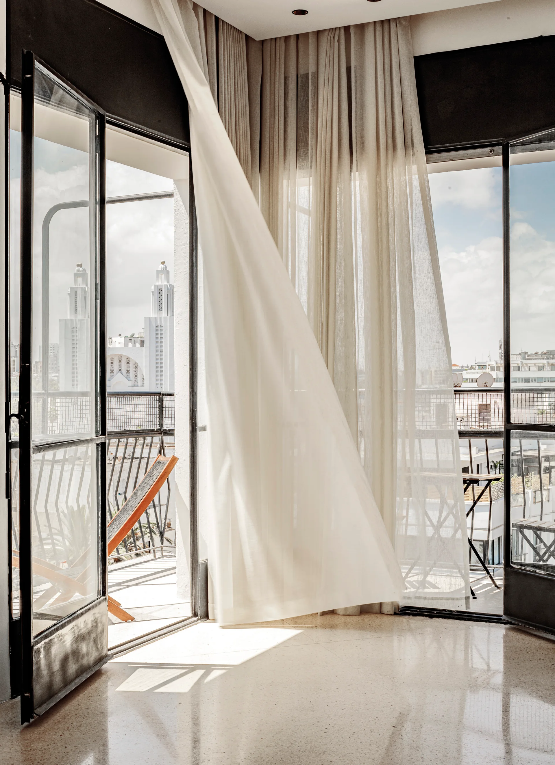An interior room looks out to a balcony through open black-framed glass doors with flowing sheer white curtains.