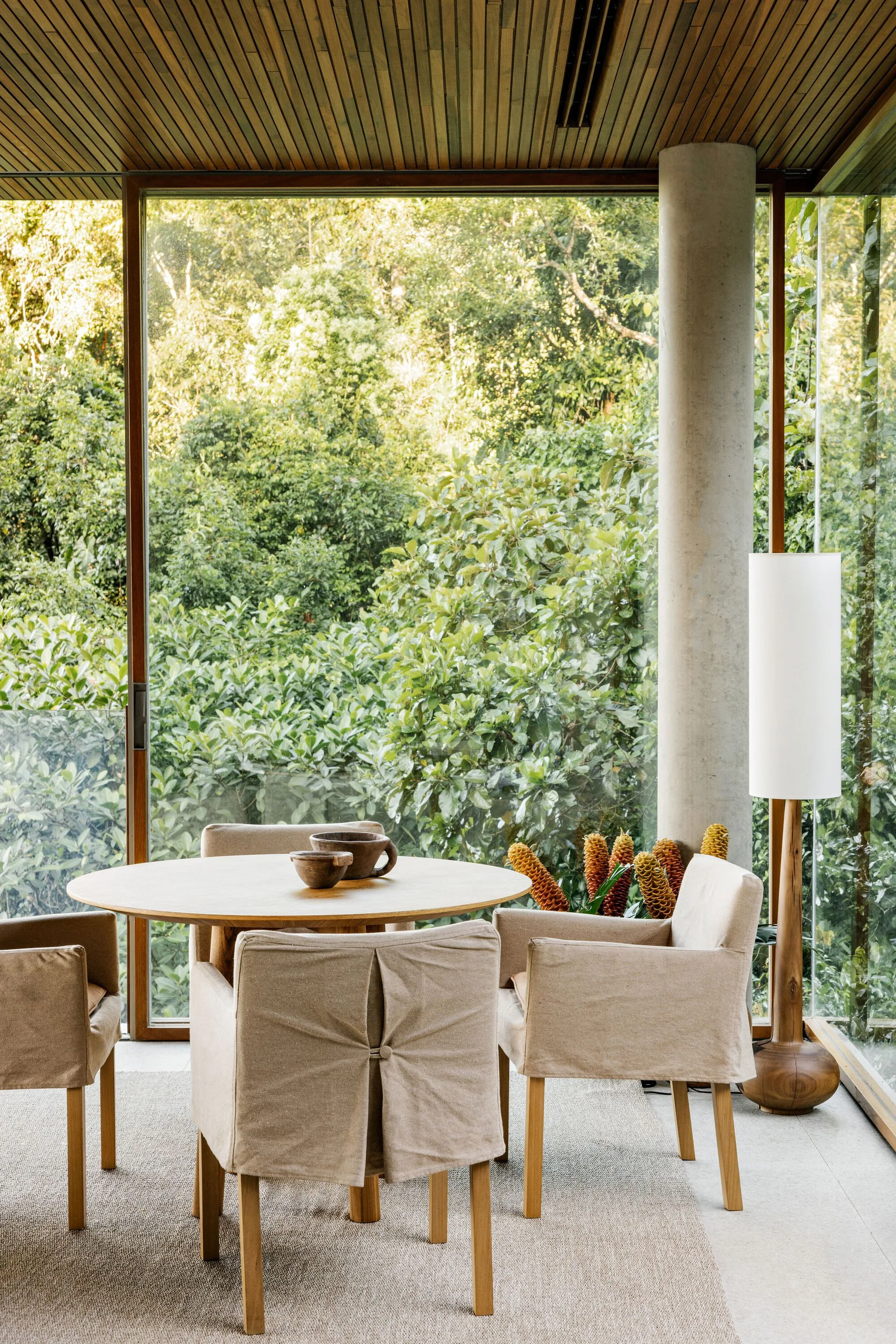 A round wooden dining table and beige slipcovered chairs sit on a neutral rug beneath a slatted wood ceiling next to large glass walls.