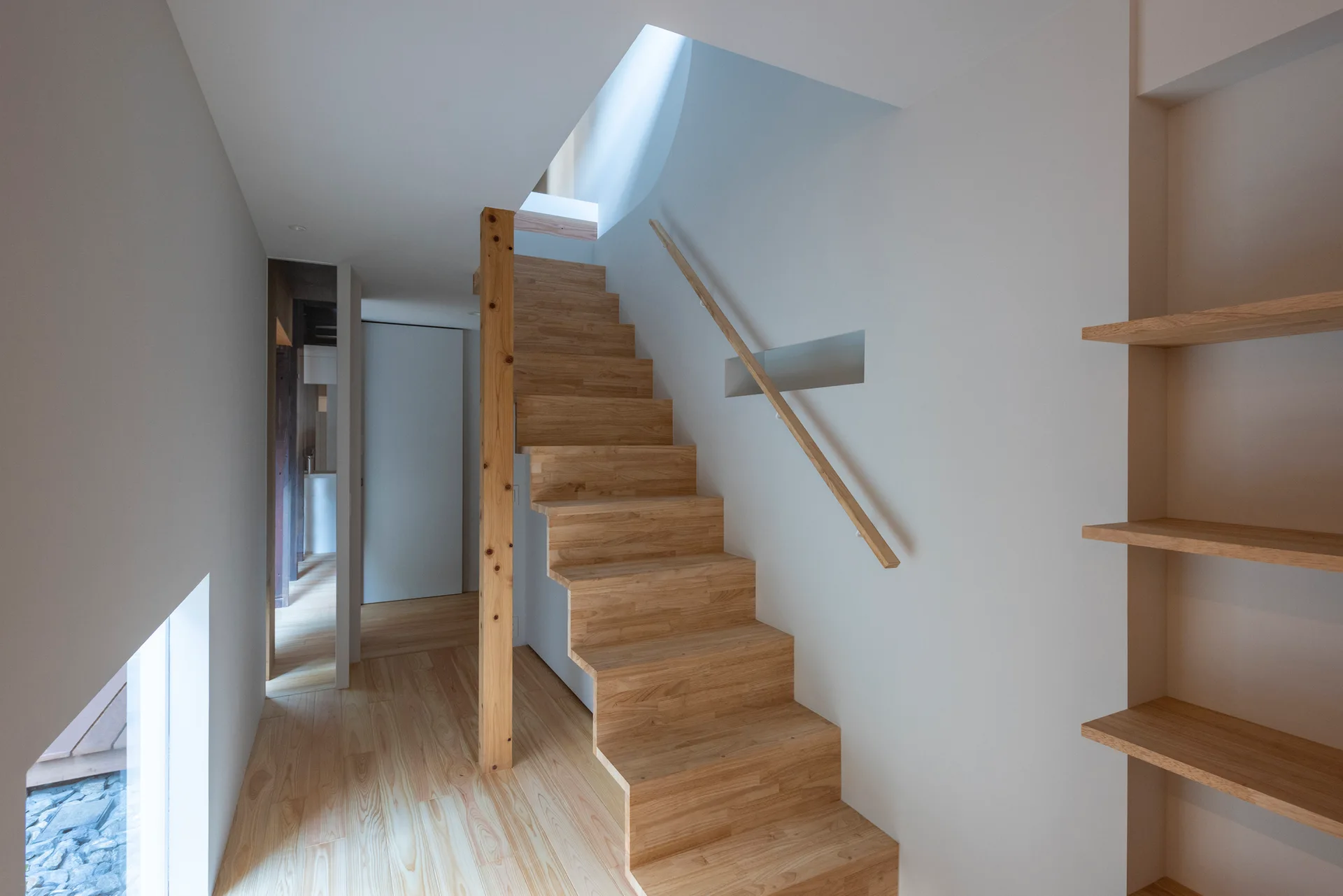 A minimalist wooden staircase with a matching handrail and built-in shelving is set against smooth white walls.