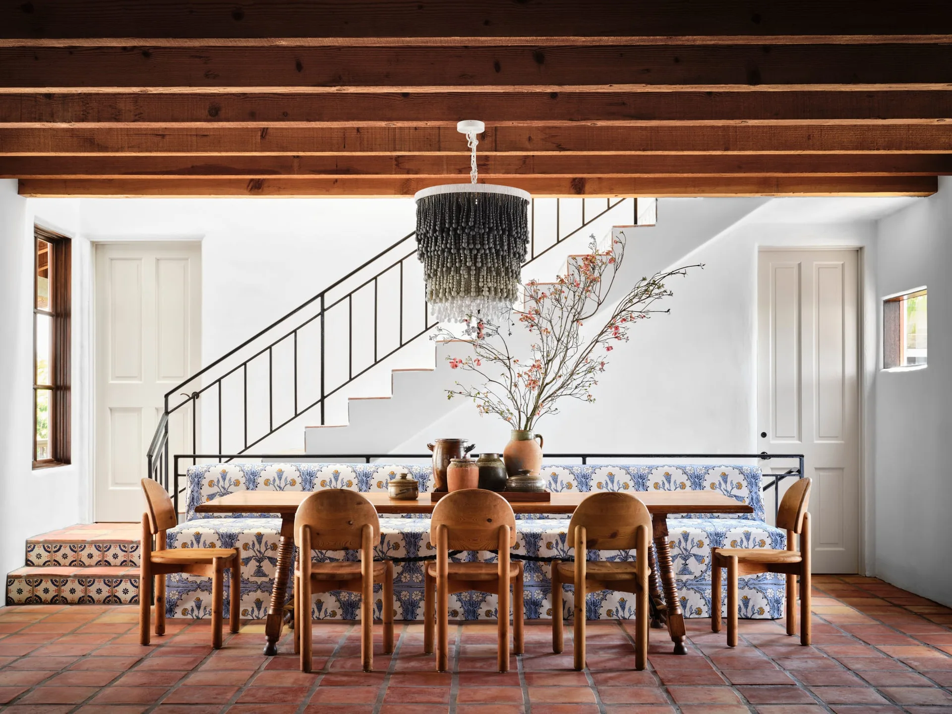 A rustic dining area features a wooden table and chairs, a patterned blue upholstered bench, a beaded chandelier, and terracotta tile flooring.