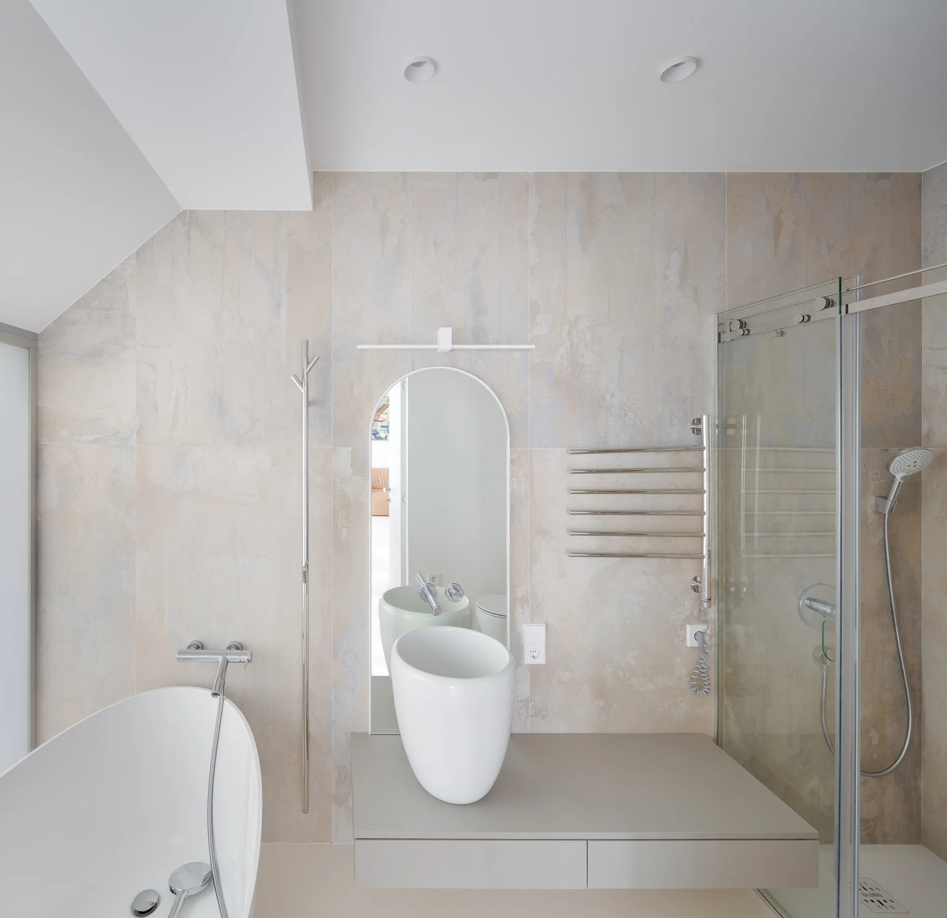 A modern bathroom with beige tiled walls, an arched wall mirror, a white vessel sink on a floating vanity, and a freestanding tub.