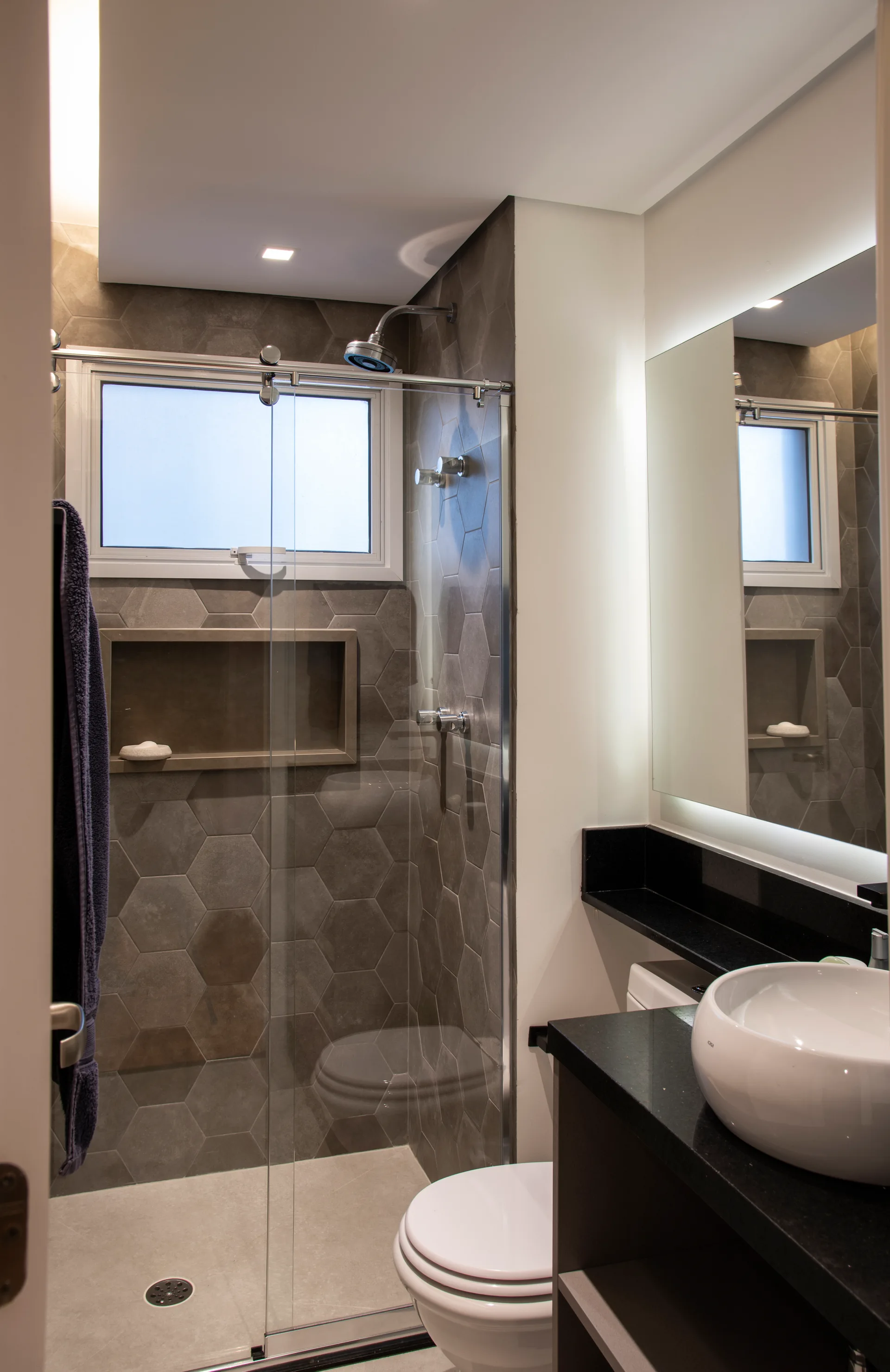 A modern bathroom features a glass-enclosed shower with grey hexagon tiles, a white toilet, and a black vanity with a vessel sink and backlit mirror.