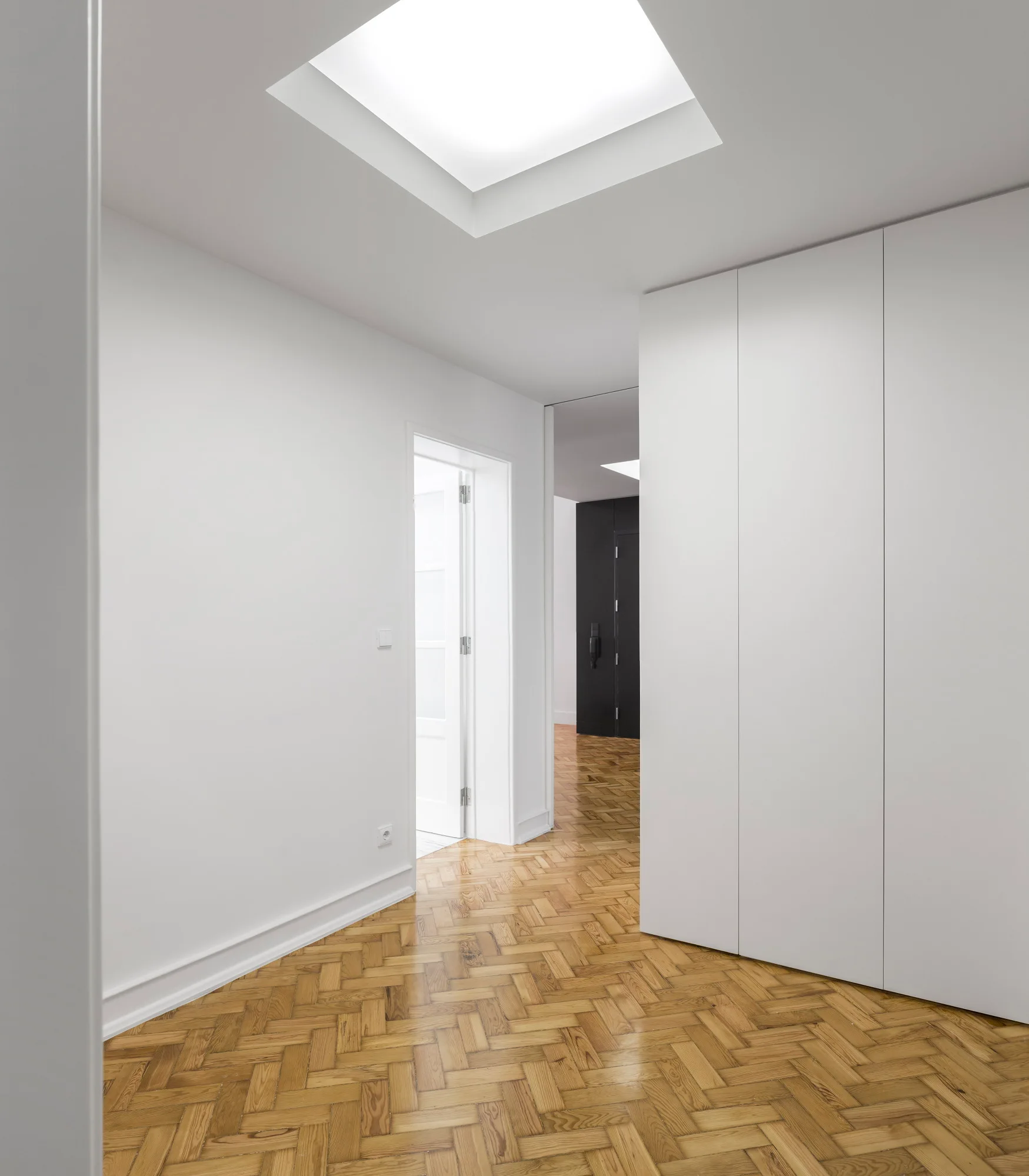 A minimalist hallway features white walls, a built-in closet system, and light brown herringbone wood flooring beneath a square skylight.