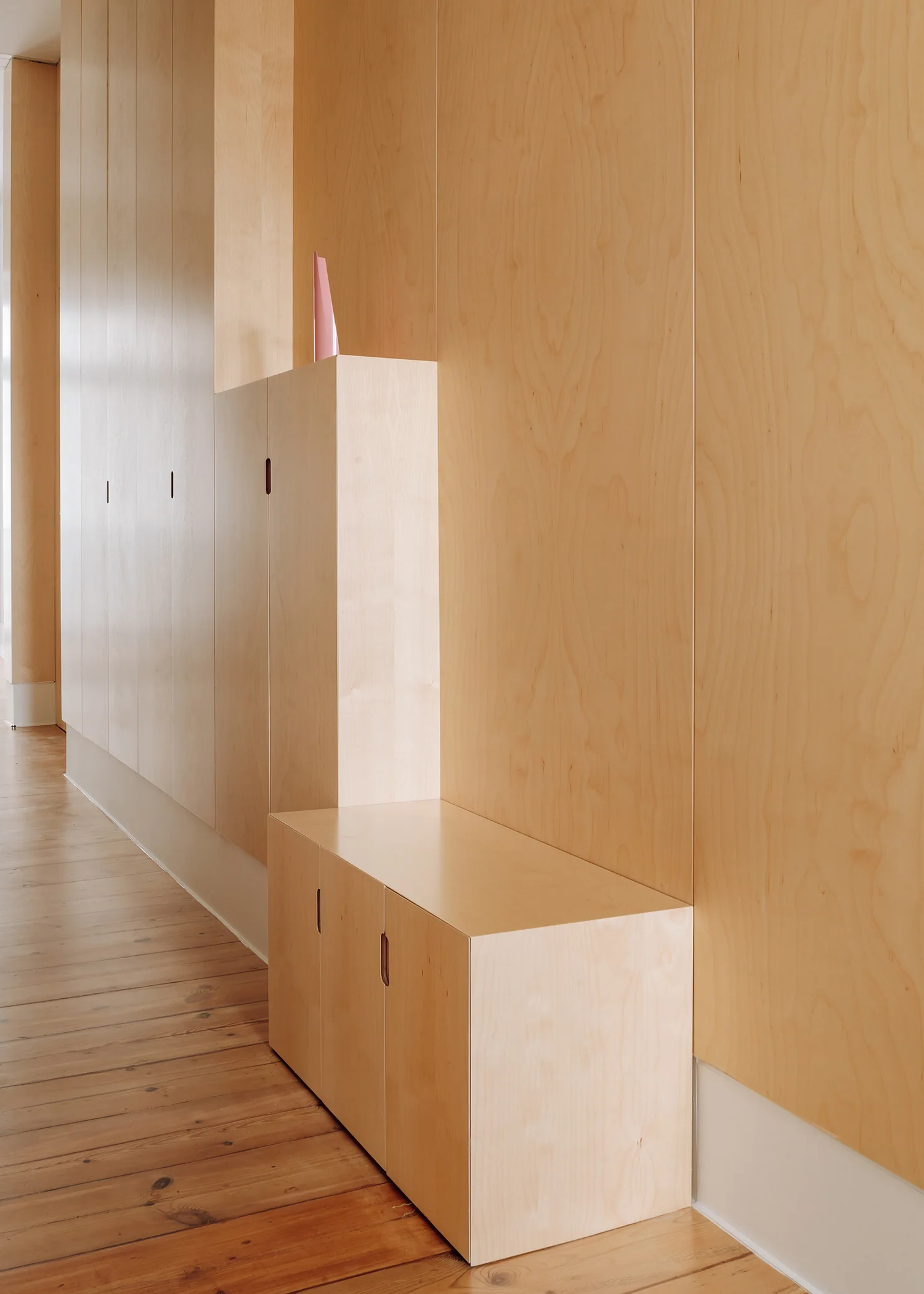 A hallway features light wood wall paneling with integrated built-in cabinets, a low matching storage bench, and natural hardwood plank floors.