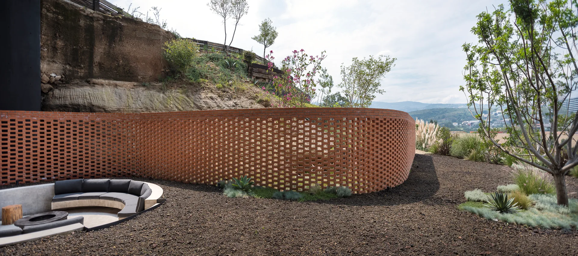 A sunken concrete patio features curved seating and a fire pit next to a long, perforated terracotta brick wall on a landscaped hillside.