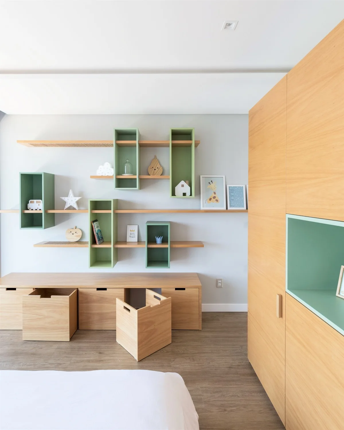 A children's bedroom featuring custom light wood built-in cabinets, mobile toy storage bins, and open wall shelves with green accents.