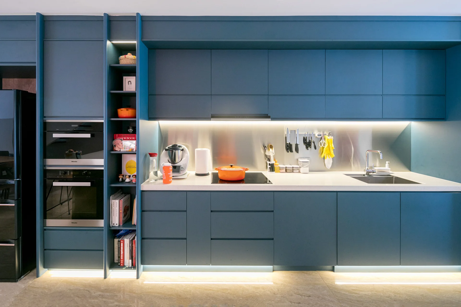 Modern kitchen featuring dark blue handleless cabinets, a metallic backsplash, white countertops illuminated by under-cabinet lighting, and built-in ovens.