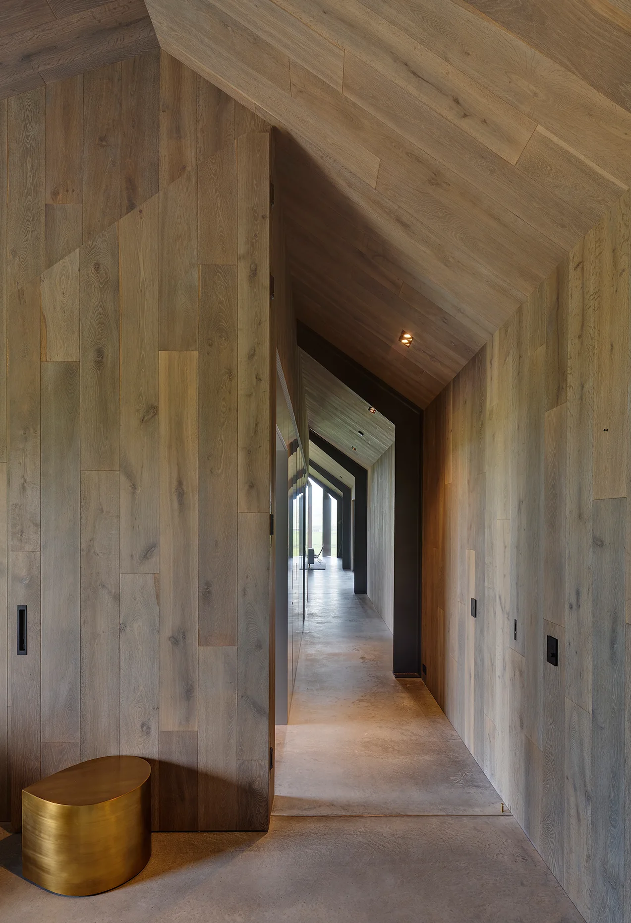 A long minimalist hallway features vertical wood plank paneling on the walls and vaulted ceiling, paired with a smooth concrete floor.