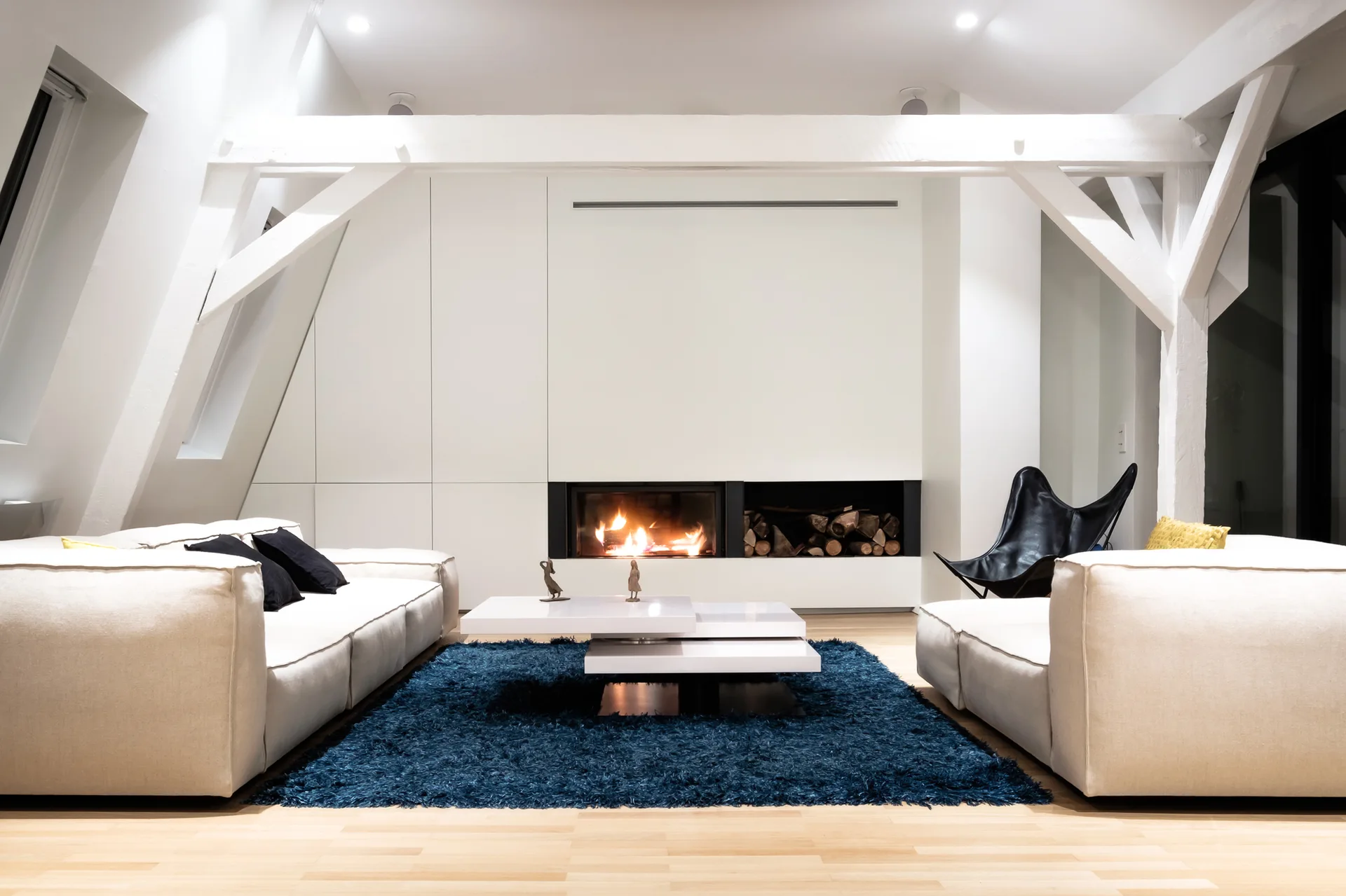 A minimalist attic living room features two white sofas, a deep blue shag rug, and a built-in fireplace under painted structural beams.