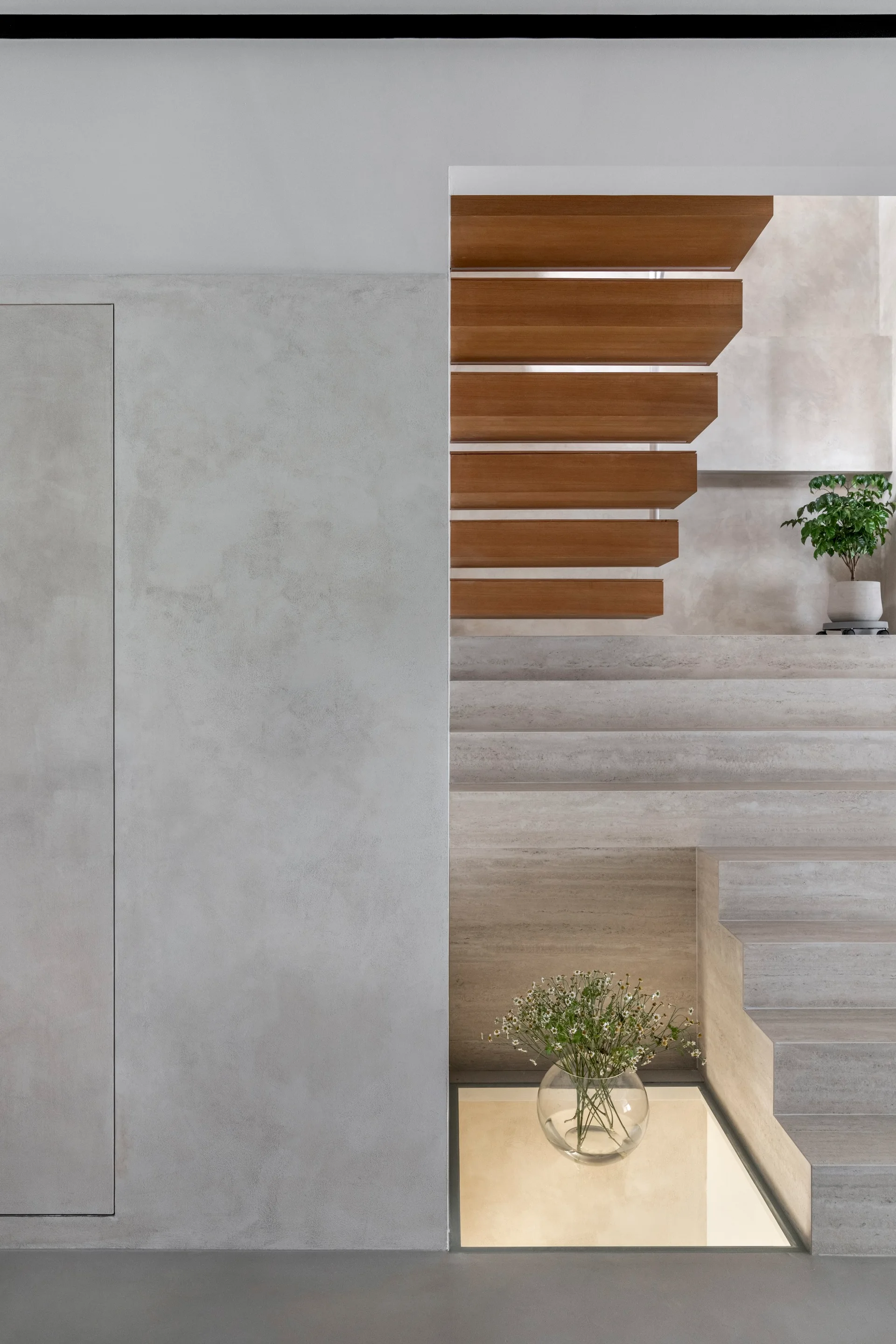 A minimalist staircase with floating wood treads above stone steps next to a grey plaster wall and an illuminated floor recess.
