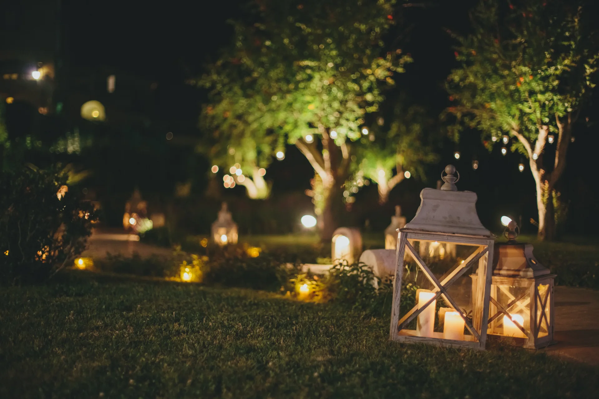Two rustic white lanterns with lit candles sit on a grassy lawn in a night garden with illuminated trees in the background.