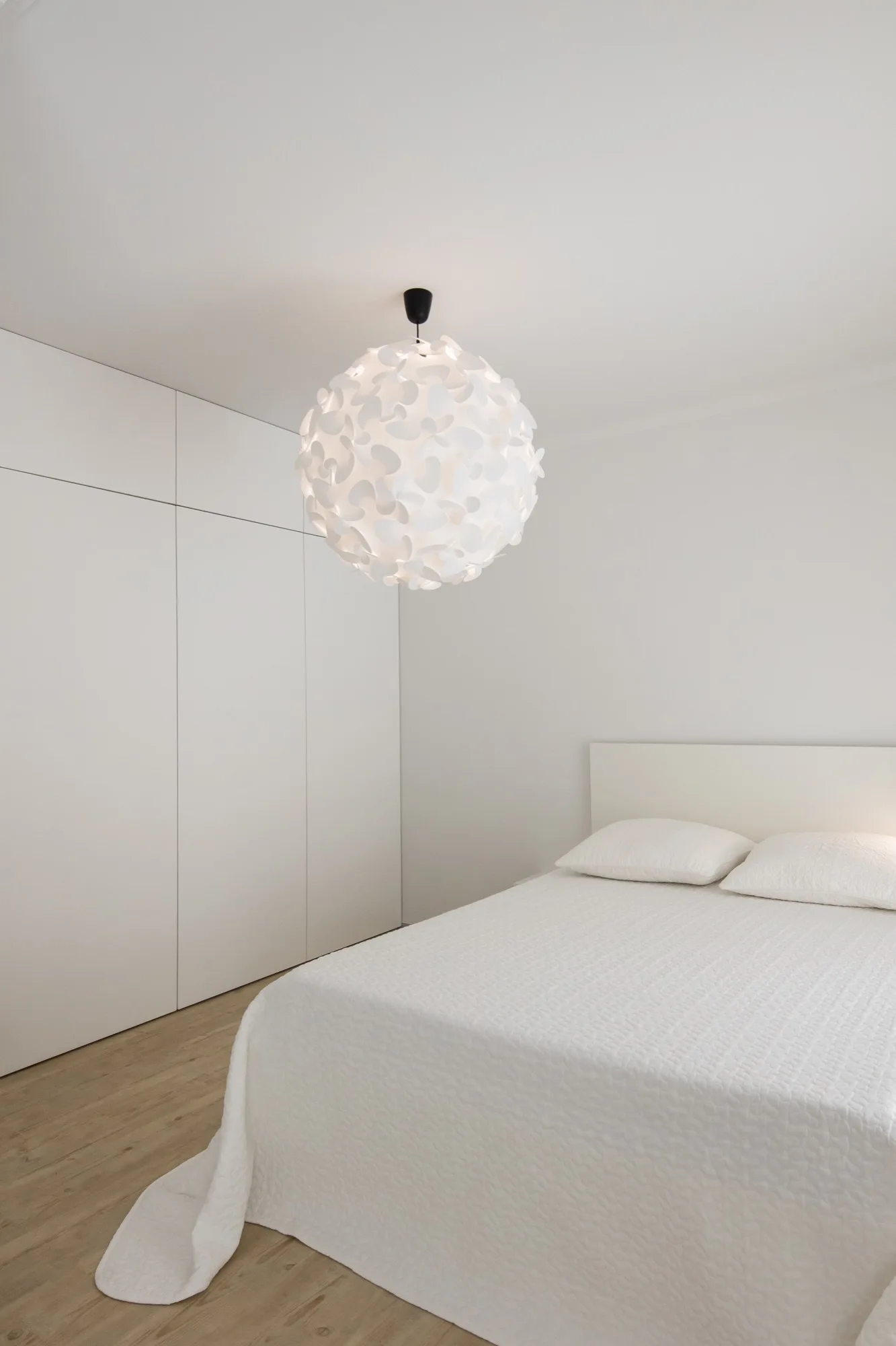 A minimalist white bedroom featuring a bed with a textured quilt, a large spherical sculptural pendant light, and a seamless built-in wardrobe.