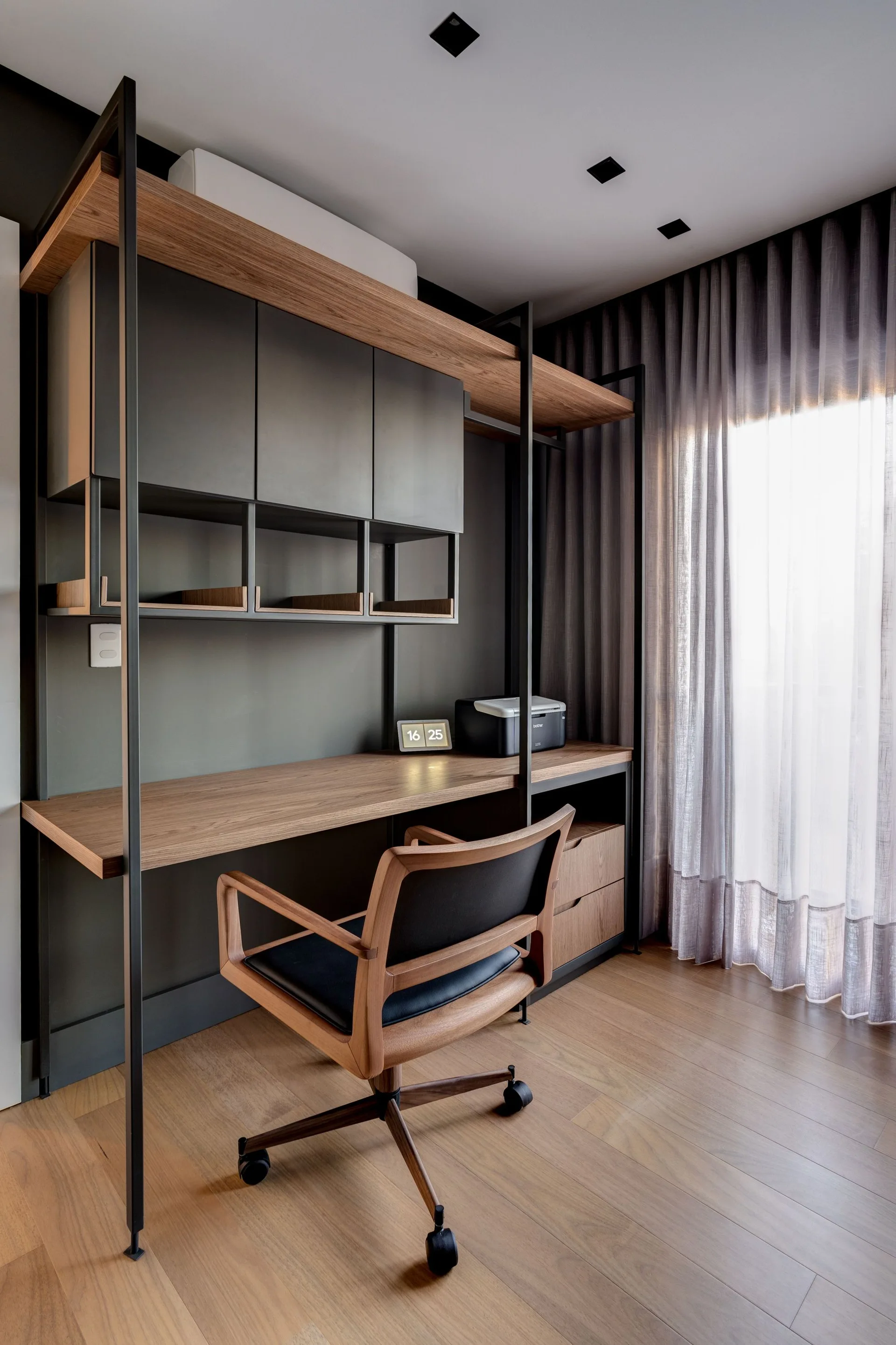 A modern home office features a built-in wooden desk with dark gray cabinetry, a matching wood desk chair, and sheer gray curtains.
