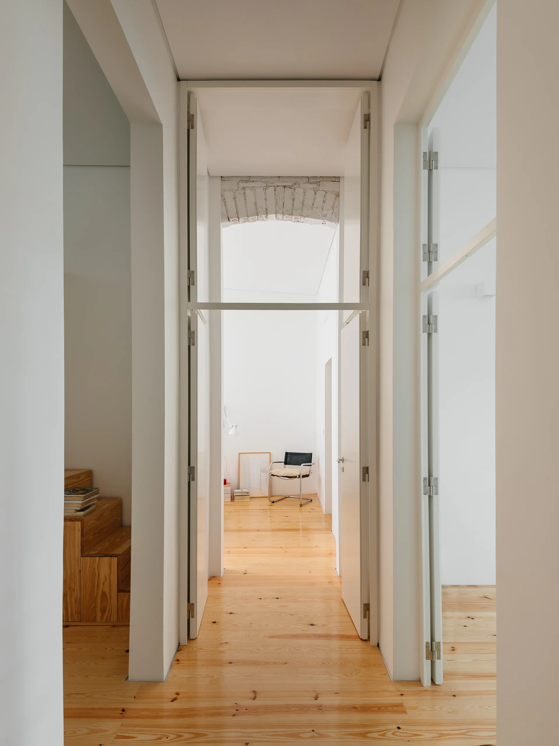 A bright minimalist hallway features light wood floors, tall white doorways, a wooden staircase, and an exposed brick arch.