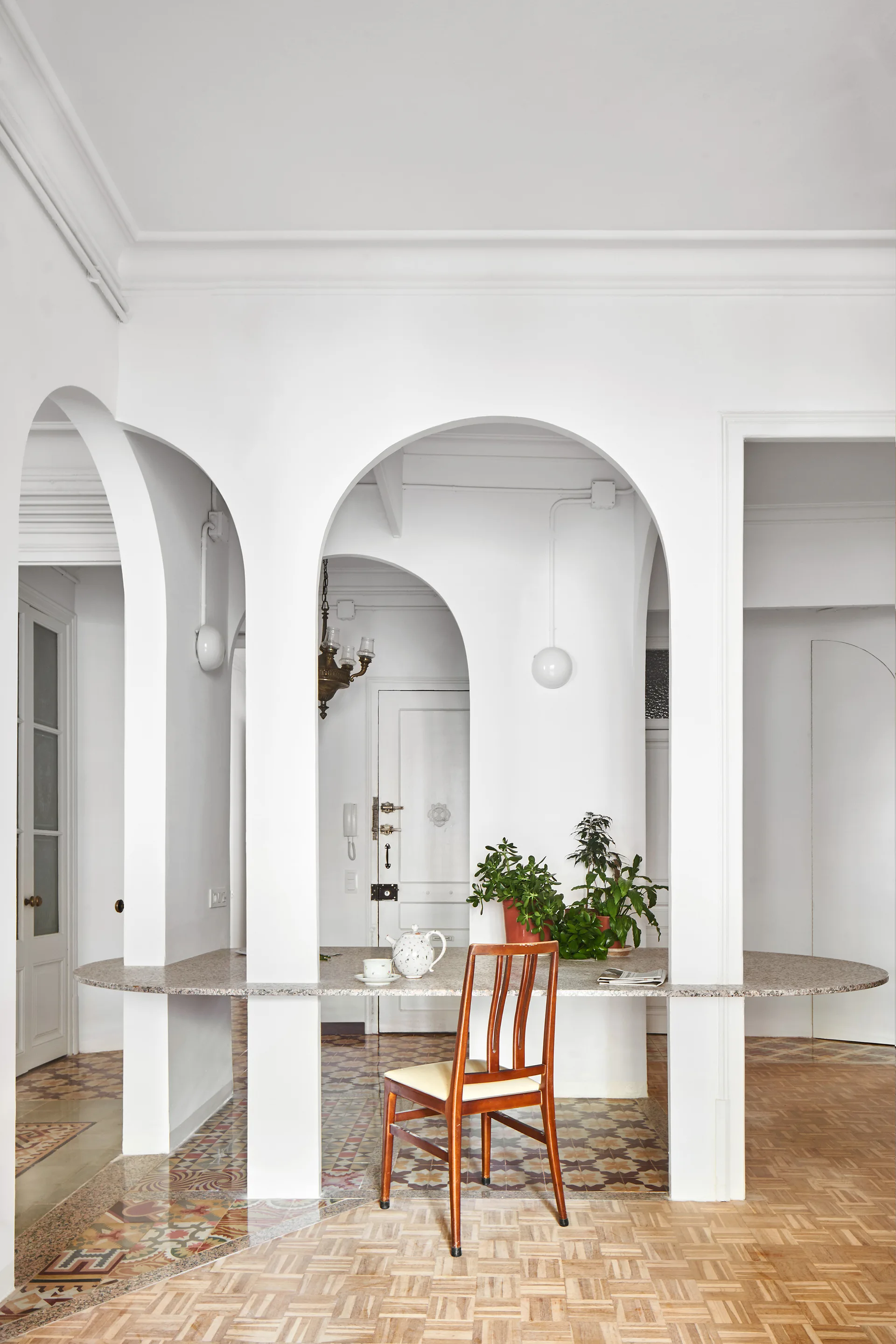 A long speckled stone dining table is integrated through white structural plaster arches, accompanied by a wooden chair and patterned geometric floor tiles.