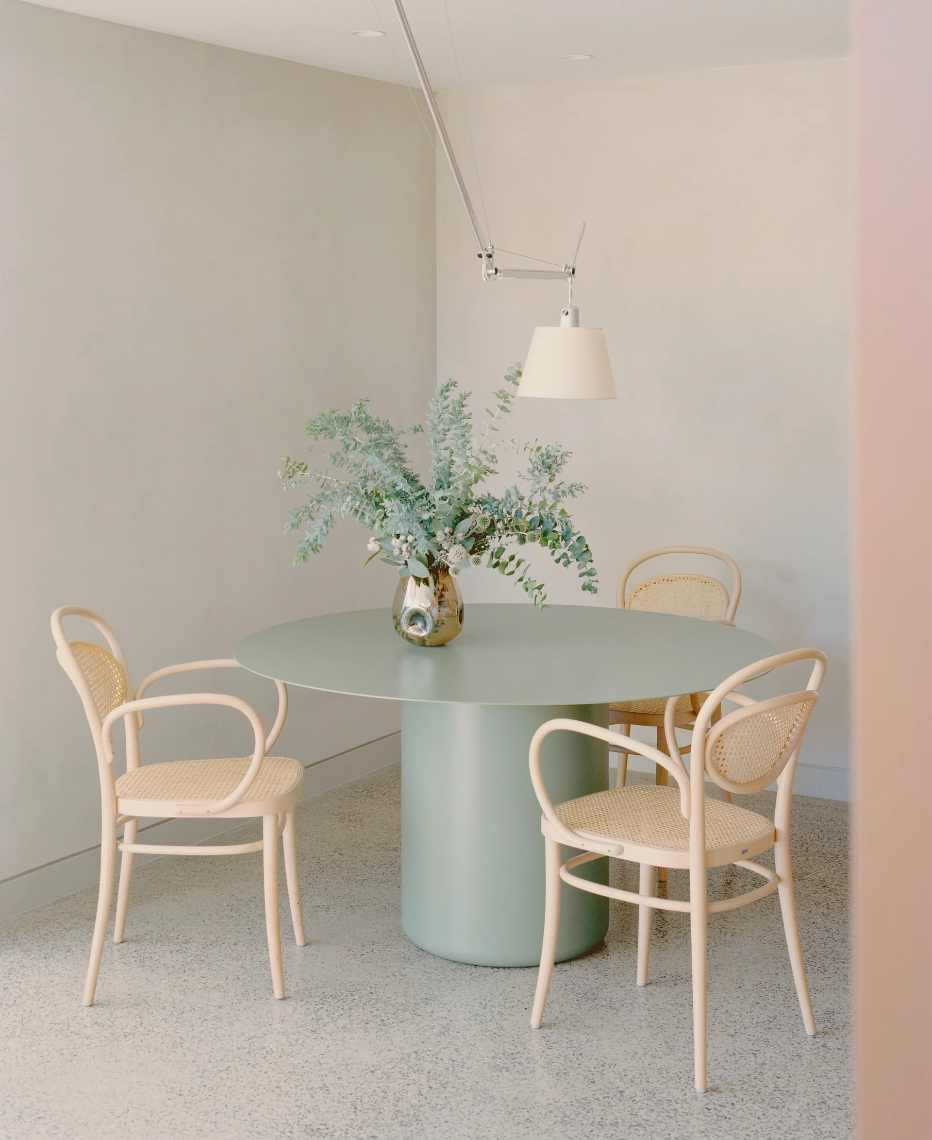A round sage green pedestal table with light wood bentwood cane chairs is set against neutral walls on a speckled stone-like floor.