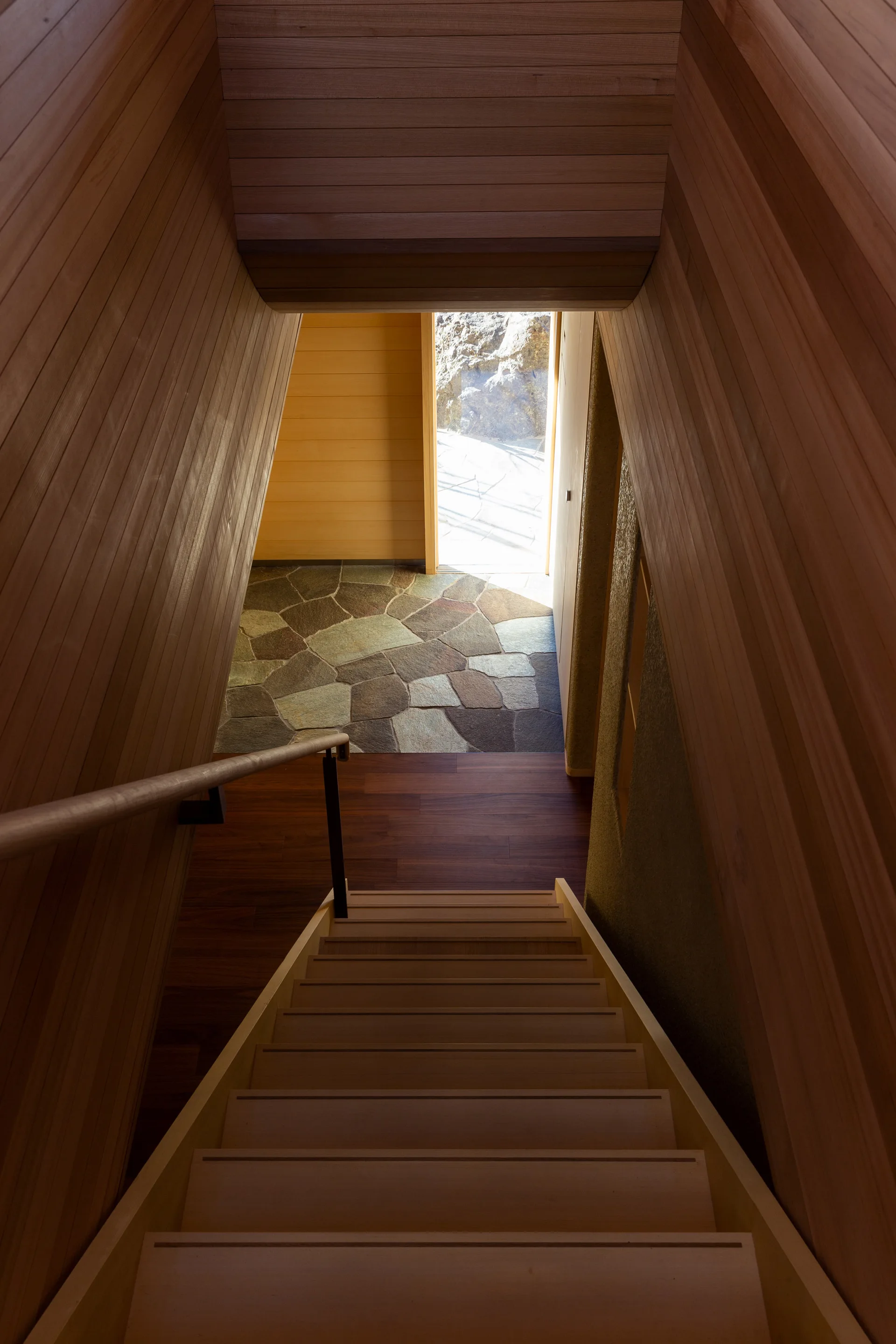 A wooden staircase with vertical wood plank walls leads down to an entryway with grey stone flooring and an open doorway letting in natural light.