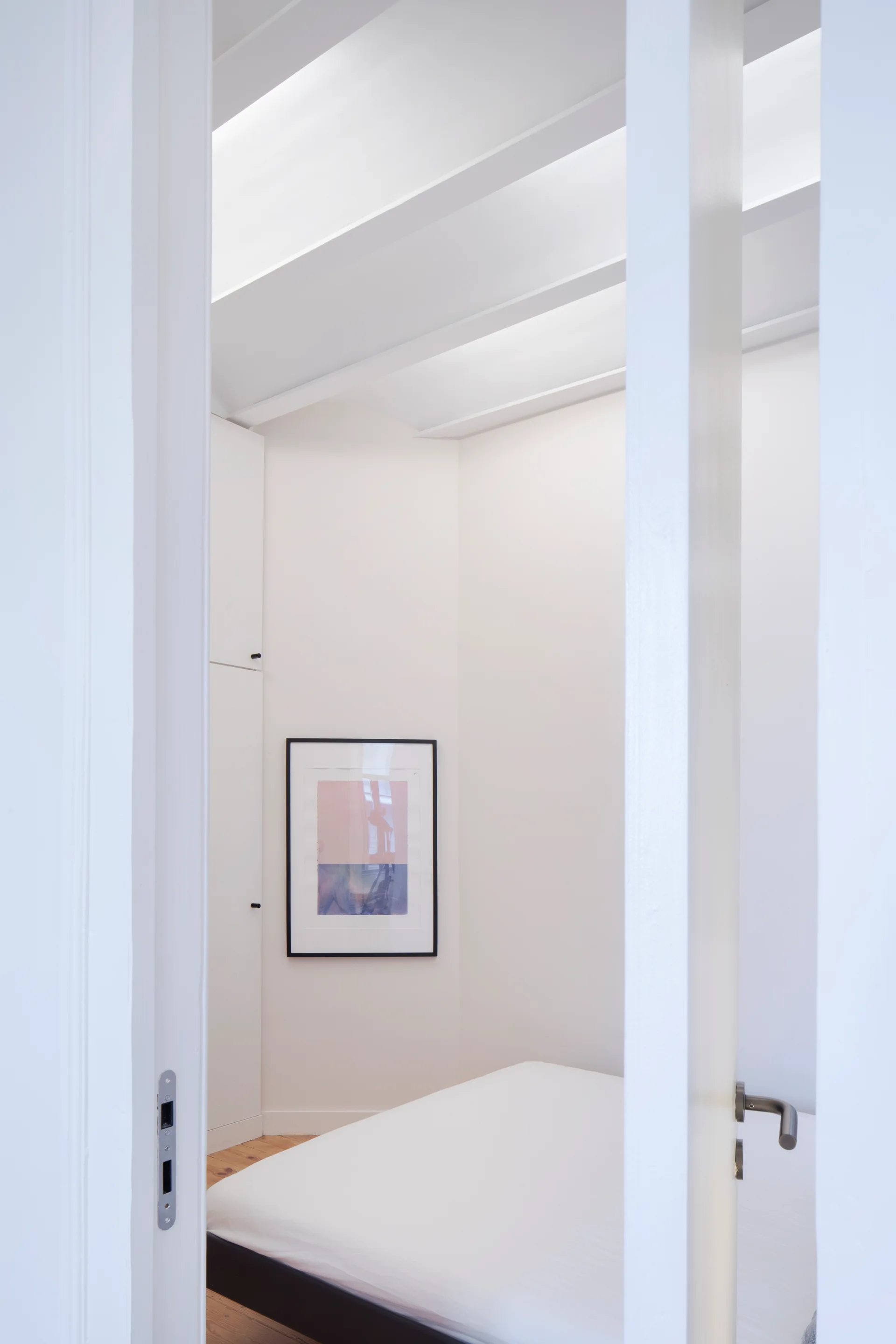 A view through a white doorway reveals a minimalist bedroom with a built-in wardrobe, a bed, exposed white ceiling beams, and framed artwork.