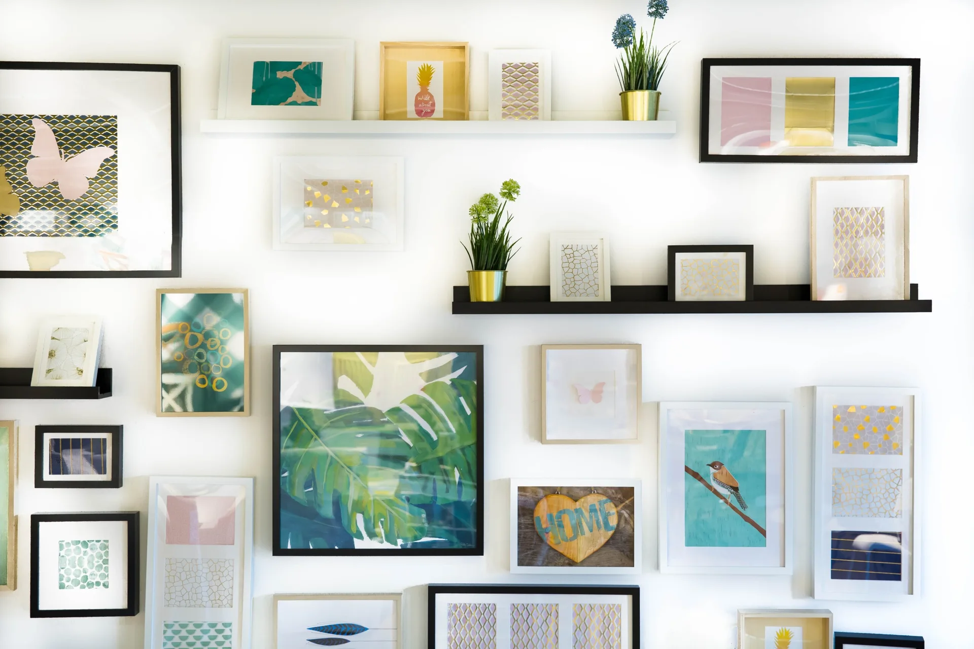 A curated gallery wall on a white background features numerous framed prints in black, white, and gold frames alongside two floating display shelves.