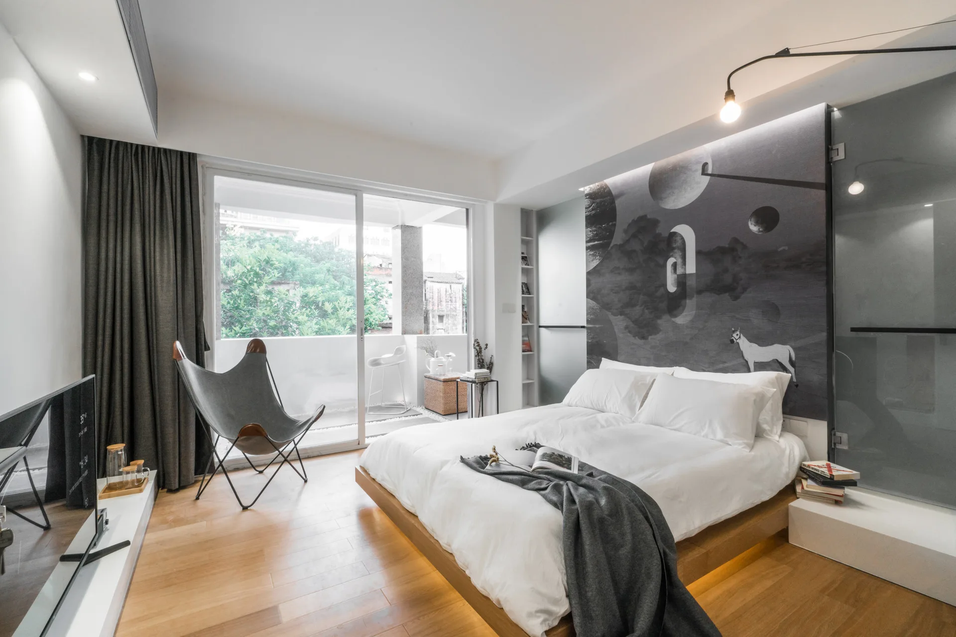 A minimalist modern bedroom featuring a low-profile wood platform bed, a large dark graphic wall mural, and a grey canvas butterfly chair.