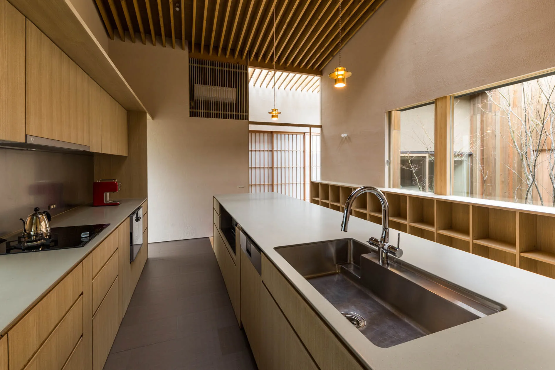 A minimalist kitchen features a long light wood island with a stainless steel sink, matching flat-panel cabinetry, and a slatted timber ceiling.
