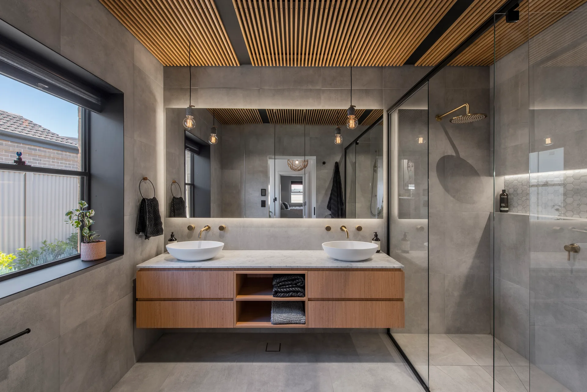 A modern bathroom with a floating wooden vanity, gray stone-look tiles, a backlit mirror, and a linear wooden slat ceiling.